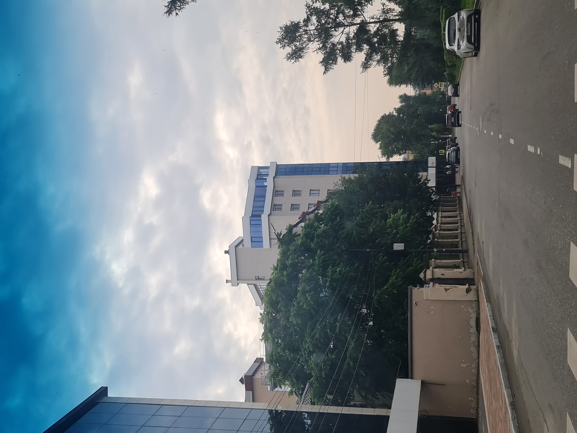 The image depicts a street scene in the city of Irkutsk, Russia, likely taken during the daytime with overcast conditions. On the left side of the street, there is a modern building with a glass facade, adjacent to a light-colored building with a wall. A large, lush green tree stands in front of the buildings, with power lines running through its branches. Further down the street, several other buildings of varying architectural styles are visible, including a tall, multi-story structure with a blue-tinted glass facade on its upper floors.
The street itself is paved with asphalt and marked with dashed white lines. Several cars are present on the road, including a silver car parked on the right side and other vehicles driving away from the viewer. The right side of the street is lined with trees, some of which appear to be pine trees. The sky is a mix of blue and white clouds, with a few small dark specks that could be birds or debris. There are no clearly identifiable people engaged in any specific actions. The overall impression is of an urban environment with a mix of modern and older architecture, surrounded by greenery.
