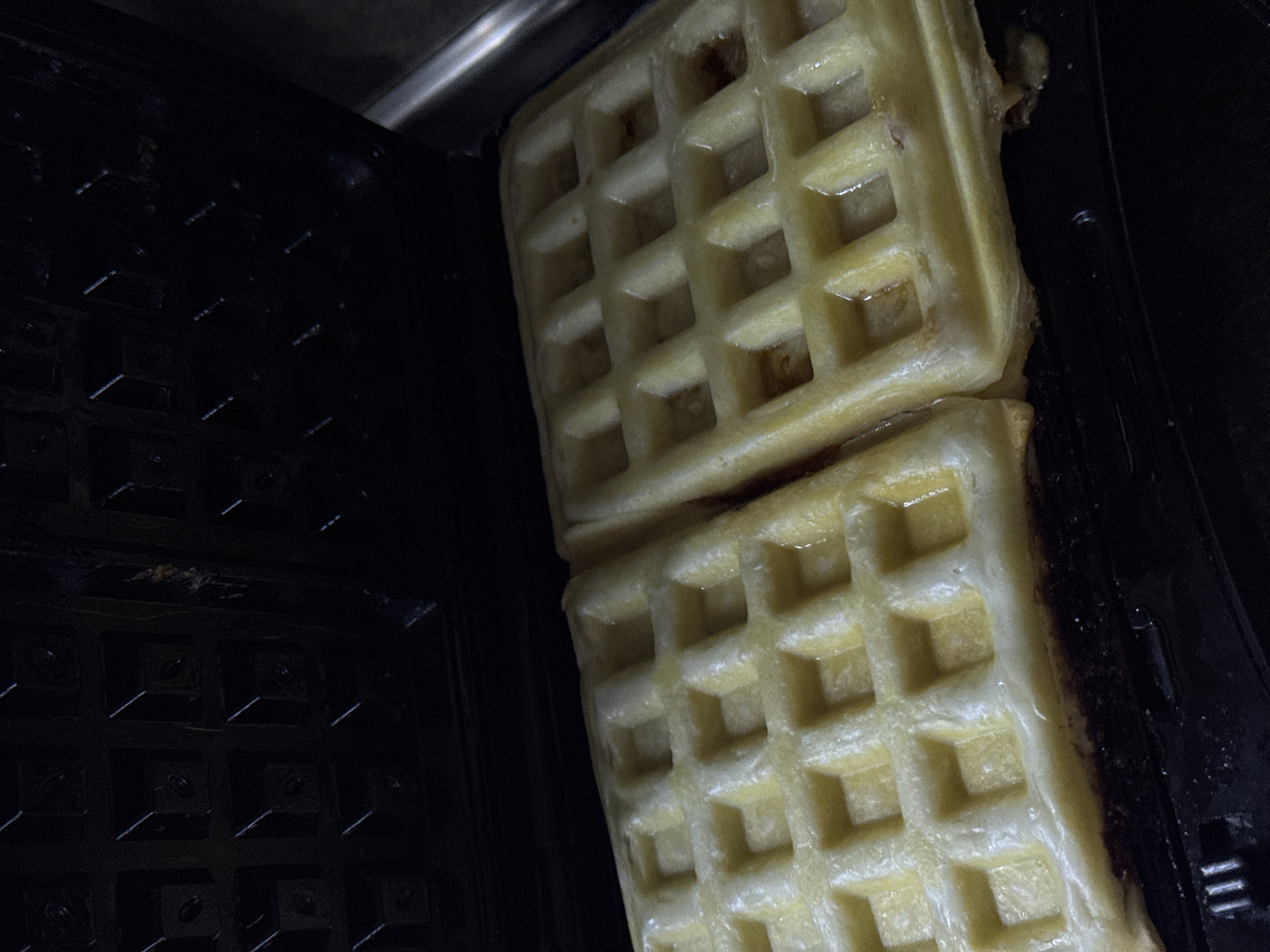 This image depicts two waffles being cooked inside an open, black electric waffle maker in an indoor setting, presumably a kitchen, in Yerevan, Armenia. The waffles are light golden-yellow, exhibiting a distinct grid pattern, and appear moist with visible sheen from cooking oil or butter. The top lid of the waffle maker is open and visible in the upper portion of the frame, showing its dark, textured cooking surface. The bottom cooking surface, also dark, holds the two waffles, with slight overflow of batter visible around their edges. No people are visible.