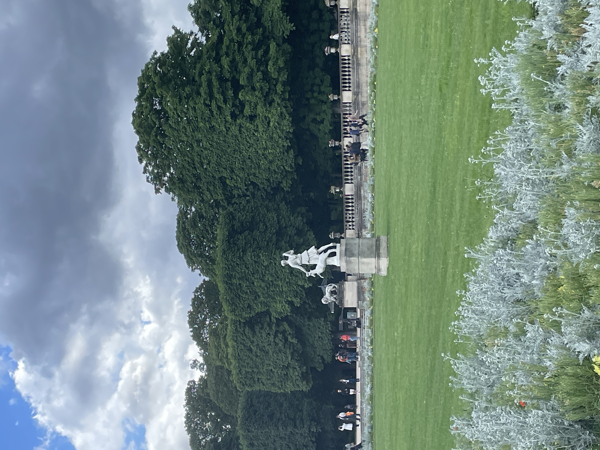 The image displays a serene park scene, likely in an urban setting, oriented as if viewed from a slightly elevated position. The dominant features include a vast, well-maintained green lawn that stretches across the middle and right side of the frame. To the left, a dense line of tall, dark green trees forms a natural backdrop, suggesting a mature arboretum or forested section of the park.
In the mid-ground, centered against the treeline, stands a prominent white statue of a male figure, possibly depicting a mythological hero or allegorical representation, mounted on a stone pedestal. To its immediate right and slightly below, there is another, smaller, darker-colored sculpture or statue.
Separating the lawn from the distant trees and statues is a decorative stone balustrade or railing, along which several people are visible. They appear to be casually walking or standing, enjoying the park environment. The balustrade suggests a defined pathway or elevated terrace.
The foreground on the right is dominated by a lush patch of silvery-green, dusty miller-like shrubs or bushes, adding texture and a contrasting color to the vibrant green of the grass. The sky above is largely cloudy, with a mix of darker grey clouds and brighter white clouds, indicating an overcast or partly cloudy day. Patches of blue sky are visible in the upper left corner. The lighting is diffused, suggesting it is daytime, perhaps mid-morning or afternoon, without harsh shadows. No discernible text is visible in the image. Given the provided context, the setting is consistent with a grand public garden in Paris, France.