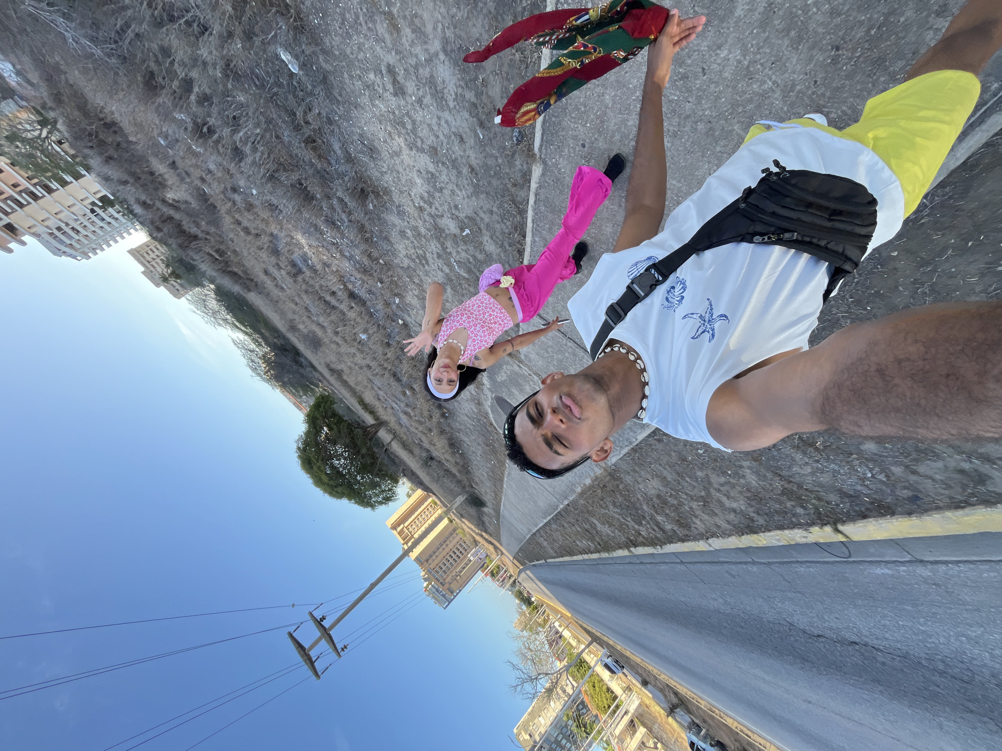 A wide-angle selfie captures two young adults lying on a paved surface outdoors, looking up at the camera. The image is significantly tilted, creating a disorienting yet dynamic perspective.
In the foreground, a male subject wears a white t-shirt adorned with blue starfish and shell graphics, bright yellow shorts, and a black fanny pack across his chest. He also sports a white shell necklace and a dark baseball cap worn backward with sunglasses resting on it. He holds up a colorful, patterned piece of fabric or scarf in his right hand. His expression is a slight pout towards the camera.
Behind him, a female subject wears a pink floral halter crop top, vibrant pink pants, and black shoes. She has a light-colored headband in her hair and a small, light-colored pouch attached to her pants. She smiles subtly at the camera, with her arms slightly spread.
The background, viewed from this unconventional angle, features a clear blue sky dominating one side. Utility poles with wires and several multi-story buildings are visible in the distance, alongside scattered trees and dry, scrubby vegetation. A road with a yellow curb is also present, suggesting an urban or suburban setting. The bright, clear blue sky and strong light indicate it is daytime. The overall impression is one of a relaxed and playful moment in a sunny outdoor environment, likely Porlamar, Venezuela. No discernible text is visible in the image.