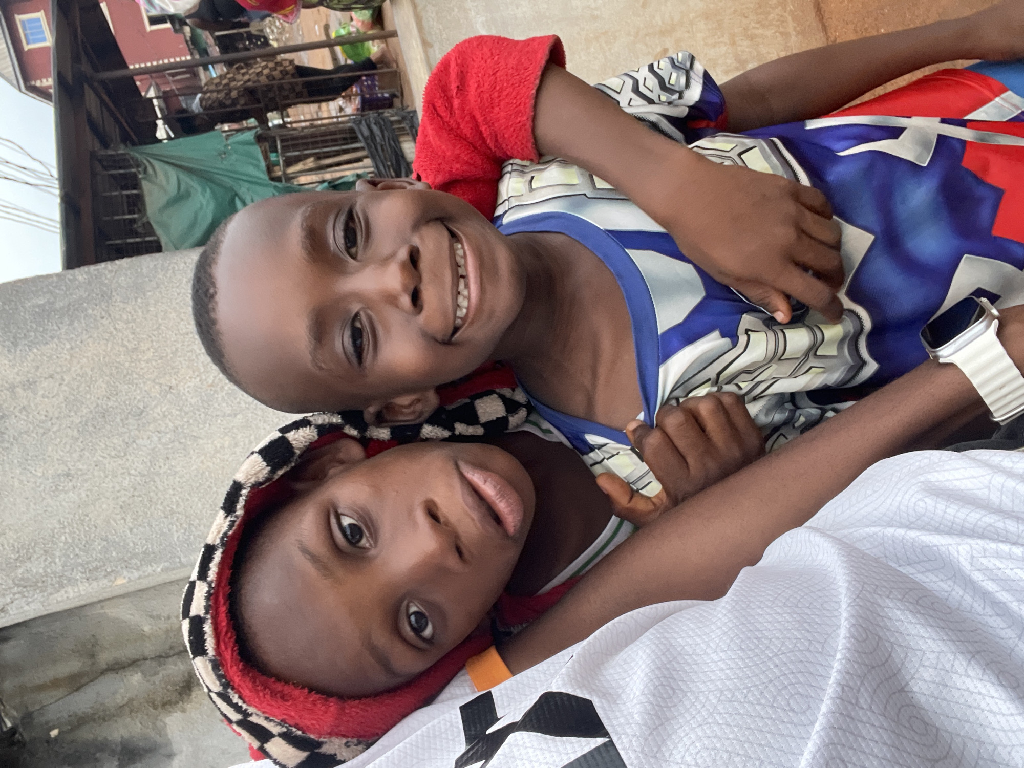 Two young boys are closely embracing and smiling at the camera. One boy wears a checkered hood and a red lining, while the other is in a blue, white, and red patterned shirt. An arm with a white smartwatch is also visible. The background shows a concrete structure and a glimpse of buildings, suggesting an urban setting in Onitsha, Nigeria.