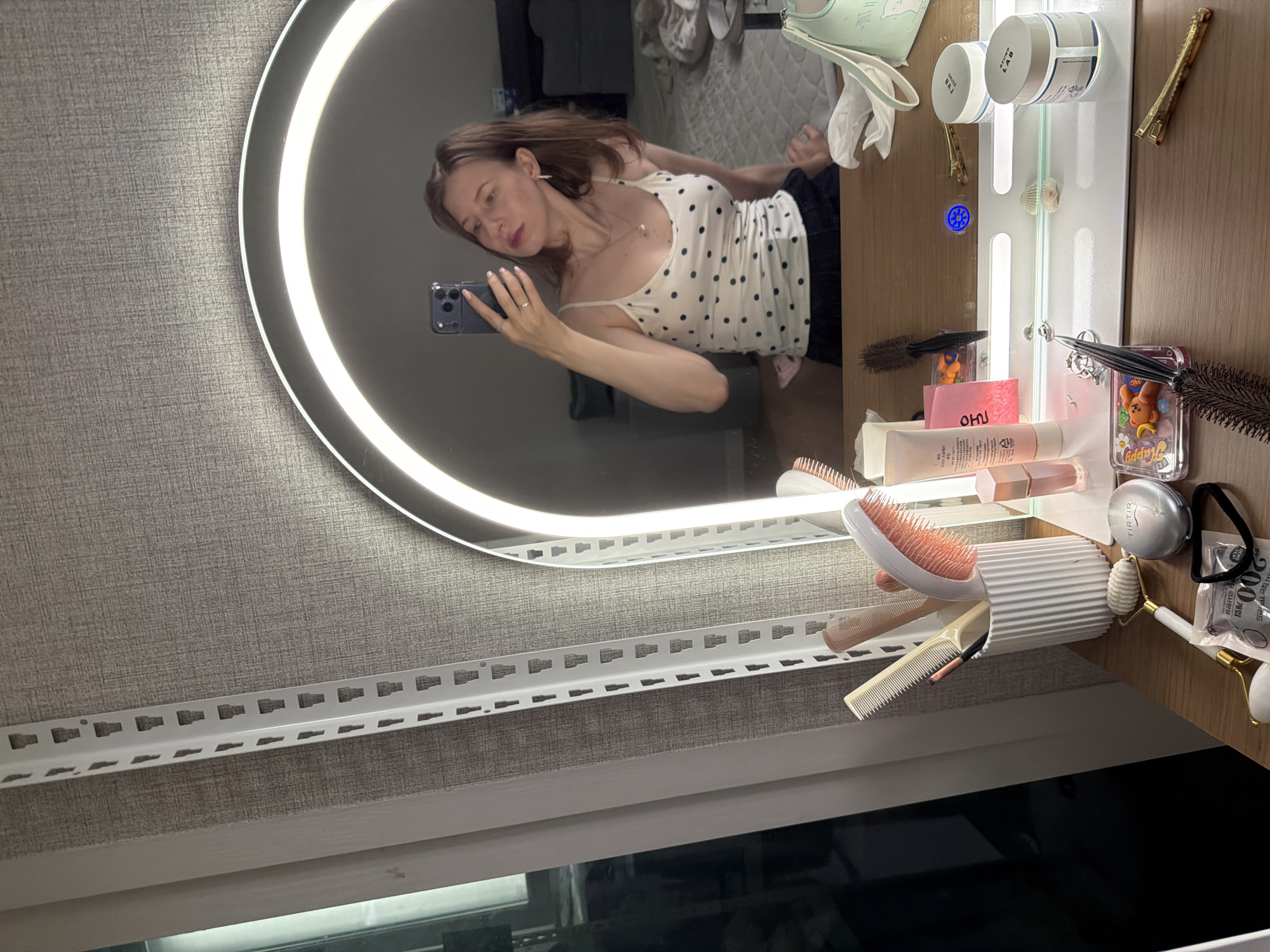 A young woman is taking a selfie in a vanity mirror with an LED light ring, reflecting her image and the items on the vanity. She is wearing a white polka dot tank top and dark bottoms. On the vanity are various beauty products, including makeup brushes, combs, skincare containers, a jade roller, and hair clips. The vanity is positioned in front of a textured wall and a tall white shelving unit. In the background, a bed is visible. The overall scene suggests a personal dressing area or bedroom. The image was taken in Dongducheon-si, South Korea.