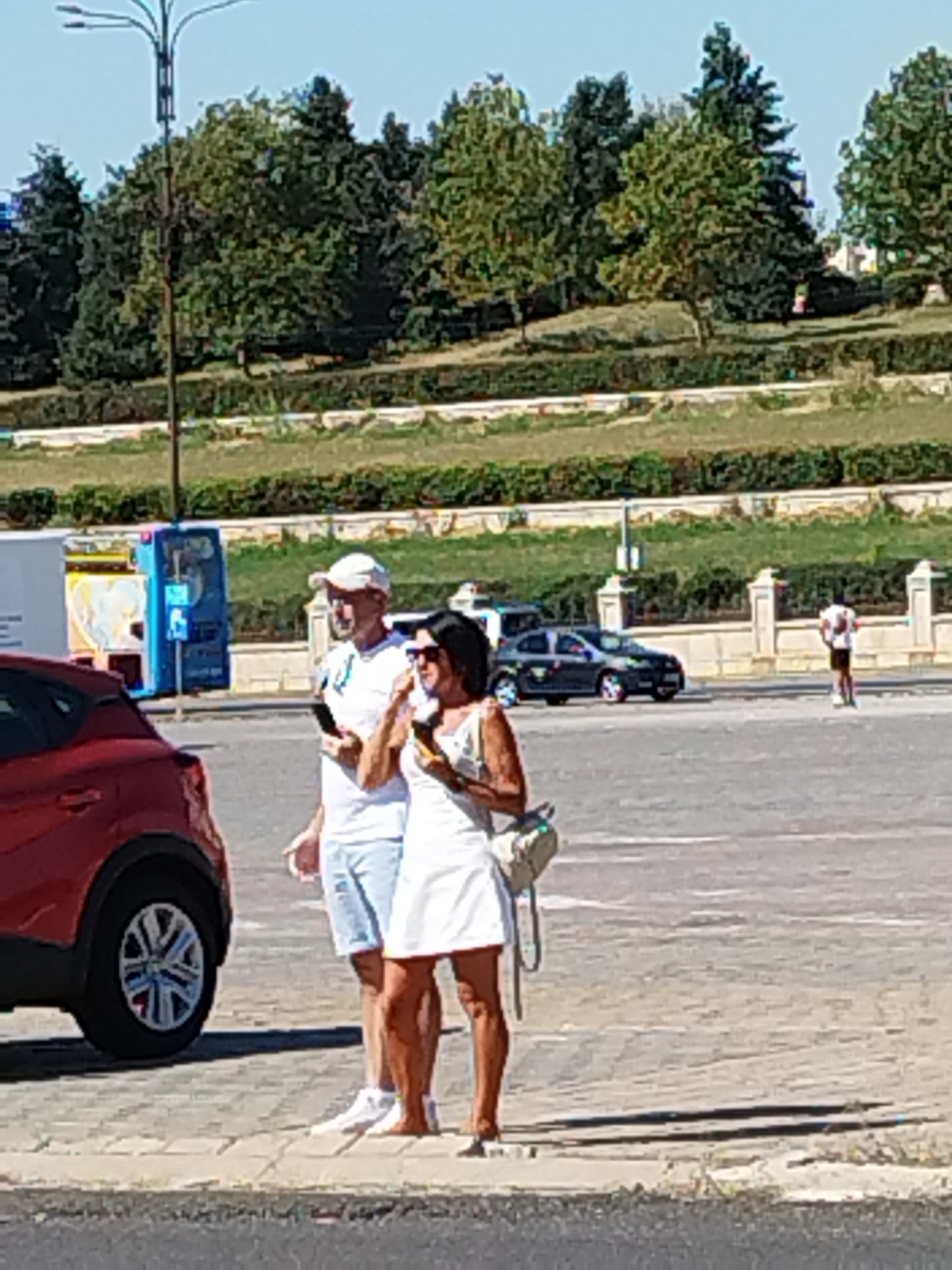 A man and a woman are standing on a paved surface in front of a red car. The man is wearing a white t-shirt, blue shorts, and a white cap, and is holding a smartphone. The woman is wearing a white dress and sunglasses, and is carrying a light-colored bag. Behind them, a dark-colored car is parked, and further in the background, a person is standing on a walkway near a fence. Trees and a slope covered in vegetation are visible in the distance, under a clear sky. The scene appears to be a parking lot in Bucharest, Romania, possibly near a park or a scenic area.