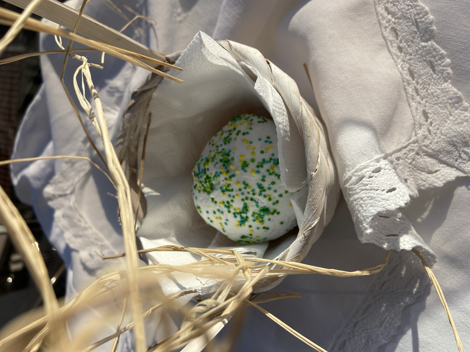 A close-up shot features a spherical, possibly confectionary item, covered in green and yellow sprinkles. It sits nestled inside a cup-like container made of layered white material, possibly dried leaves or petals, which rests upon a white fabric with a lace edge. Dry, straw-like stems are arranged around the cup. The scene is bathed in bright sunlight, casting shadows that emphasize the textures of the materials. The location in Budva, Montenegro, cannot be determined from the image.