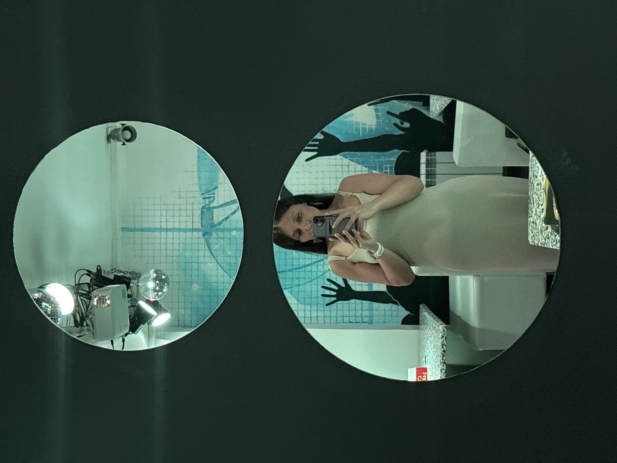 The image shows a woman reflected in a round mirror, likely taking a selfie with her phone. The mirror is mounted on a dark, matte background. The reflection includes part of her body and face. The background visible in the mirror appears to be a bathroom or similar space, with a mural of hand silhouettes on a tiled wall, a sink, and decorative elements. Another round mirror is visible to the left with disco balls and stage lights. It is presumed that the image was taken in Chaikovsky, Russia.