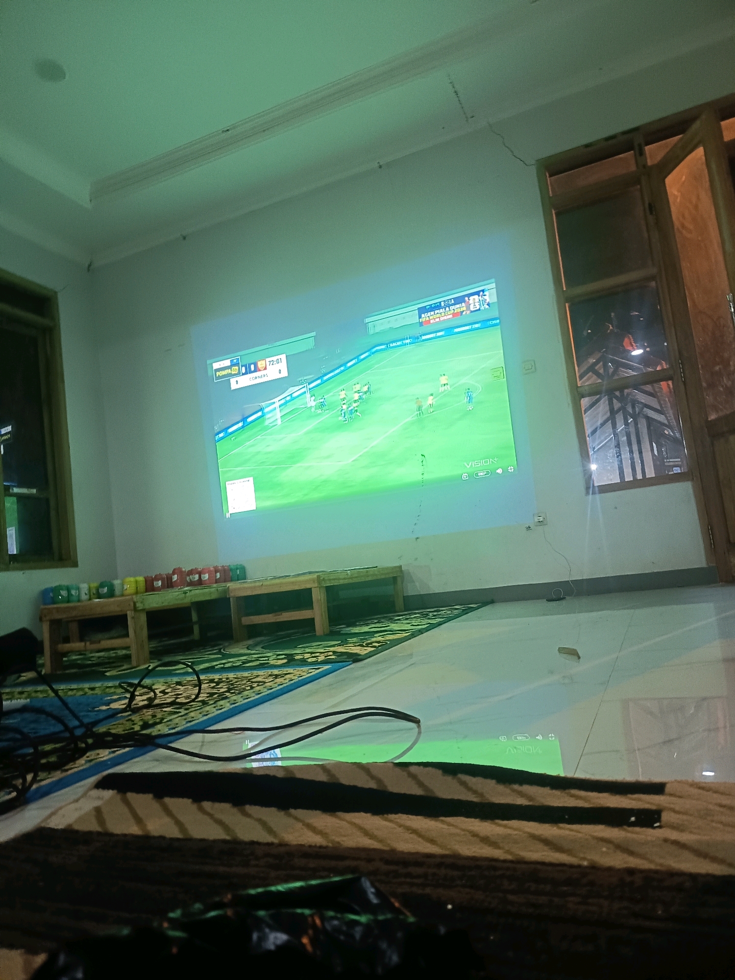 This is an eye-level, medium shot capturing a living room scene in Sumedang, Indonesia, where a projector is displaying a soccer match on a white wall. The projector, a VISION brand device, is positioned on the floor, casting the image of a game with a score display showing "72:01" and "CORNERS, 0". Two wooden benches are placed in front of the projected screen, with an assortment of colorful cups, possibly beverages, arranged on the bench closer to the screen. The floor is covered by a patterned rug, and several black cables are visible, suggesting an electronic setup. The room is dimly lit, with the projector's light illuminating the main screen and a portion of the floor. To the right, a wooden framed window with glass panes reveals a dark outdoor scene with artificial lighting visible. The wall on the left has a window with a dark pane. The ceiling has a decorative molding and a visible crack.