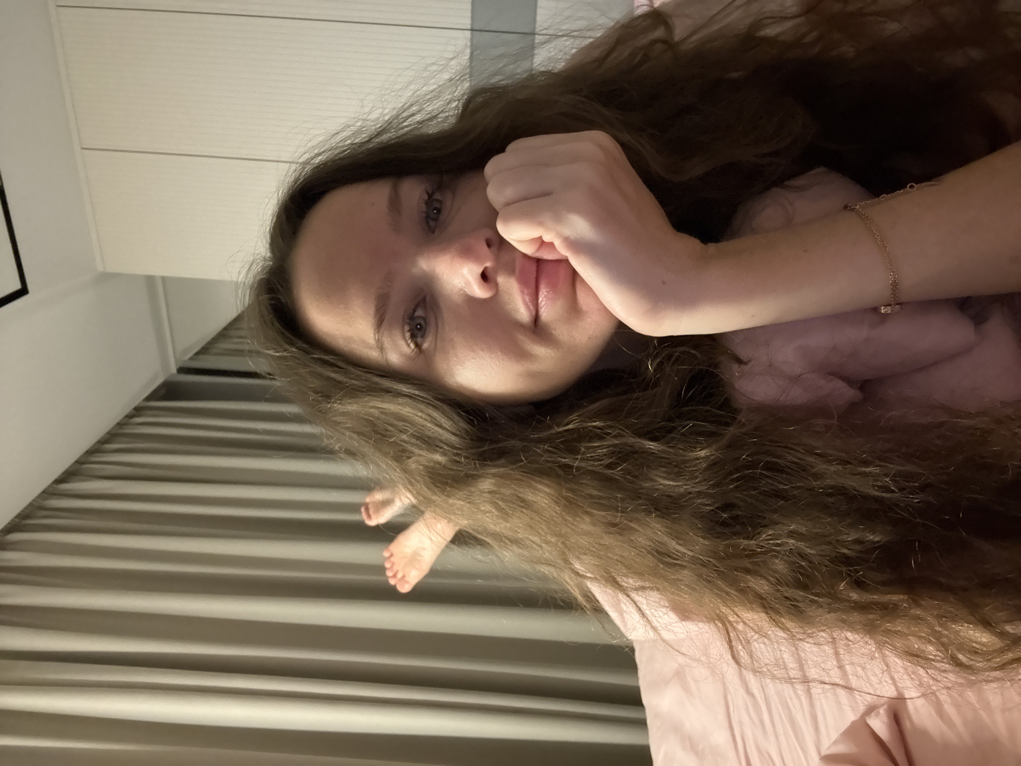 An indoor portrait shows a young adult female with long, wavy brown hair lying on a pink bed. Her right arm is bent, with her hand resting on her chin, and she is looking towards the camera. Her left leg is bent, with her foot visible, positioned in front of beige curtains in the background. The room appears to be a bedroom, with a closet visible to the right. The image was taken in Khimki, Russia.