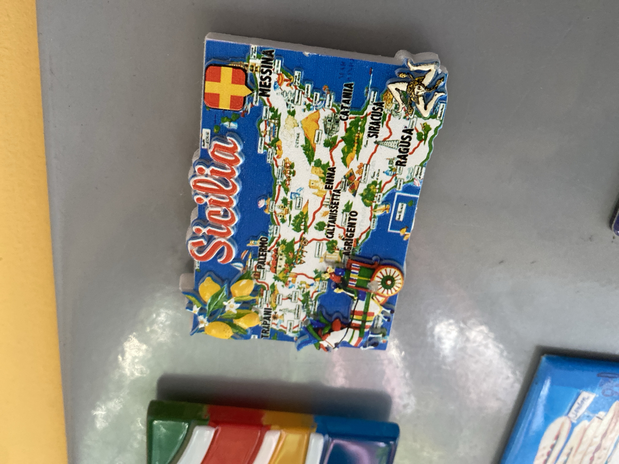 A close-up, eye-level shot shows a blue refrigerator magnet shaped like the island of Sicily. The magnet is detailed, featuring a map of Sicily with various cities labeled in black text, including Palermo, Messina, Catania, Siracusa, Ragusa, Enna, Caltanissetta, and Trapani. The magnet is decorated with small illustrations of lemons and flowers at the top, a horse-drawn cart with figures in the lower left, and Mount Etna depicted as a yellow volcano in the center. The flag of Sicily, a triskelion with a gorgon's head in the center, is also present on the magnet. The magnet is attached to a gray refrigerator, which has a yellow surface above it. To the left of the Sicily magnet is a portion of another magnet, also rectangular, with vertical stripes of red, white, and green. The background is primarily the gray surface of the refrigerator. The overall scene is static and well-lit. The location of this scene is not determinable from the image.
