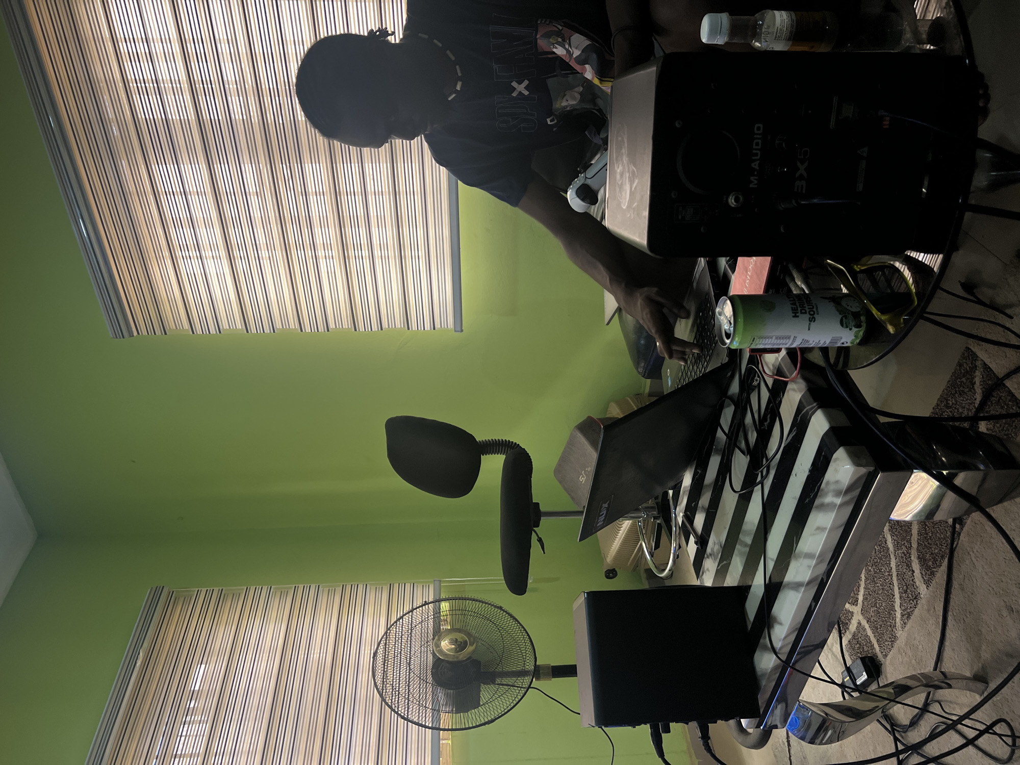A person with dark skin and dreadlocks, wearing a dark T-shirt with a graphic, is working on a laptop. The scene appears to be inside a room with green walls and blinds covering the window. A black office chair with a curved back and headrest is visible. There's a black desk fan and audio equipment including an M-Audio BX5 studio monitor. Other items on the desk include bottles, a can of what looks like a "Healthy Drinks Sours" beverage, and sunglasses. The table has a black and white stripe pattern. Given the mention of the city of Abuja, Nigeria, it can be inferred that this scene takes place indoors in Abuja.