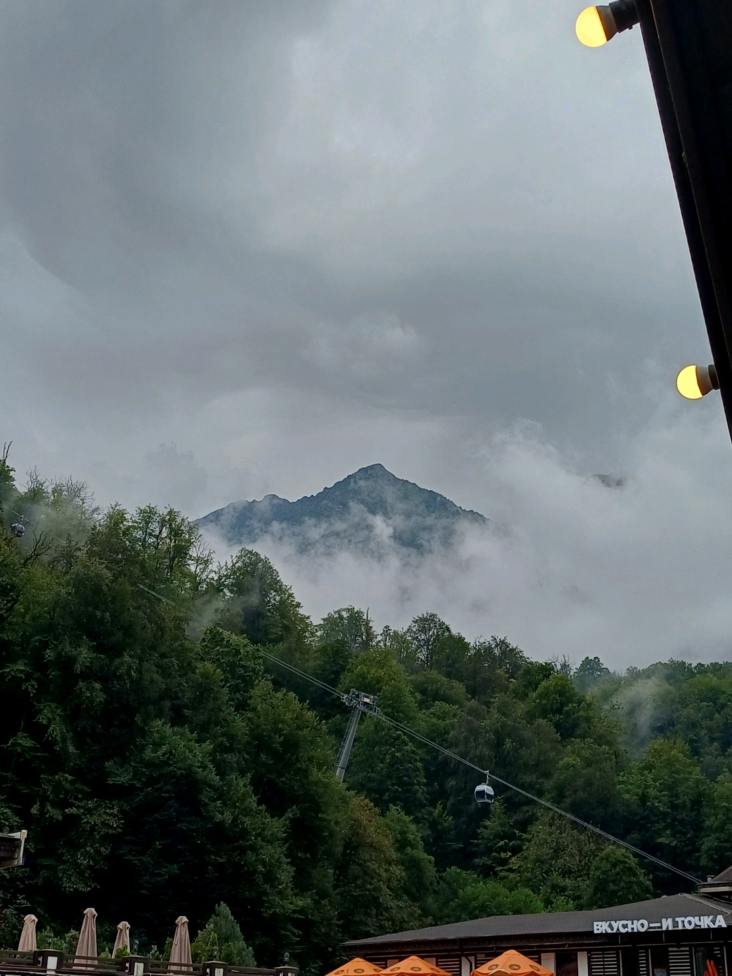 The image depicts a mountainous landscape in Estosadok, Russia, under a cloudy sky. A prominent, rugged mountain peak emerges from a layer of mist and clouds in the distance. The foreground is dominated by a dense forest of green trees covering a steep slope. A cable car with a small cabin is visible traversing the slope, suspended from cables. Below the forest, on the right side of the frame, a building with signage that reads "ВКУСНО — И ТОЧКА" (which translates to "Tasty — and that's it") is partially visible. Some umbrellas and outdoor seating arrangements are also present in the lower part of the image, suggesting a commercial or dining establishment. Two illuminated light bulbs are attached to a dark vertical structure on the right edge of the frame, one positioned higher than the other. The overall atmosphere is overcast and slightly hazy, indicating possibly cool or damp weather.
