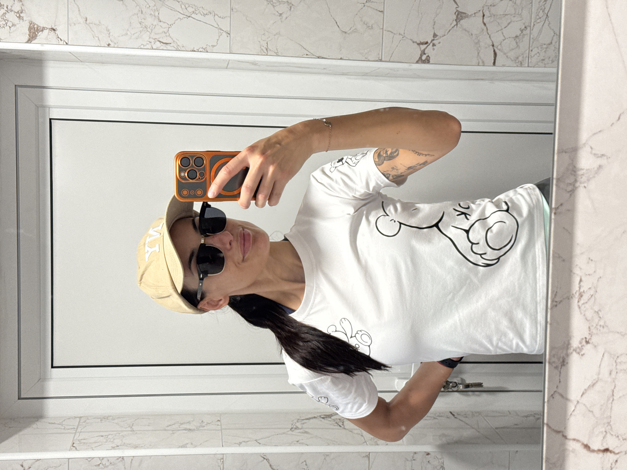 The image depicts a person taking a selfie in a bathroom. The person is wearing a light-colored baseball cap, sunglasses, and a white t-shirt with a cartoon graphic. They are holding a smartphone with an orange case. The background shows a white door or wall with a white frame and marble-patterned tiles. The location is not evident from the image. There is no information to suggest the location is in Pleven, Bulgaria.
