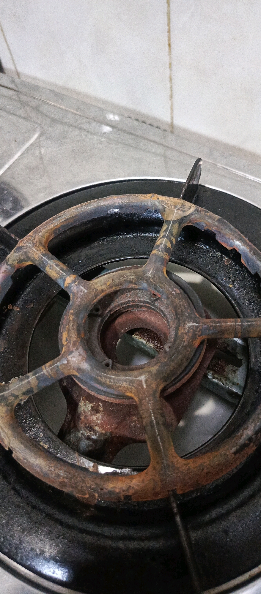 A close-up, high-angle shot shows a heavily rusted burner grate of a gas stove. The grate is made of dark metal, with thick layers of rust visible on its spokes and rim. The grate is positioned over the burner itself, which is also partially obscured by rust and grime. The surrounding stovetop appears to be made of stainless steel, with a tiled backsplash visible in the upper portion of the image. The scene is lit by overhead lighting, casting subtle shadows. There are no people or active interactions depicted. This image appears to be a photograph of a kitchen appliance in a residential setting, possibly located in Sidoarjo, Indonesia, given the context provided.