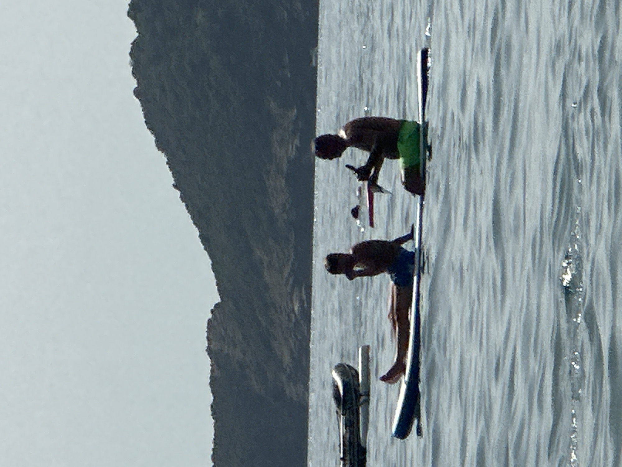 In Boreti, Montenegro, two individuals are on stand-up paddleboards in a body of water with a hilly, tree-covered landmass in the background. On the left, a person sits on a paddleboard with their legs extended. They are wearing dark shorts and their torso is bare. To their right, another person is kneeling on a paddleboard, facing the viewer's left. This individual is wearing green shorts and is bare-chested, holding an object that appears to be a paddle. A small inflatable boat is visible to the left of the first paddleboarder, resting on the water. The water is calm with gentle ripples. The sky is overcast.