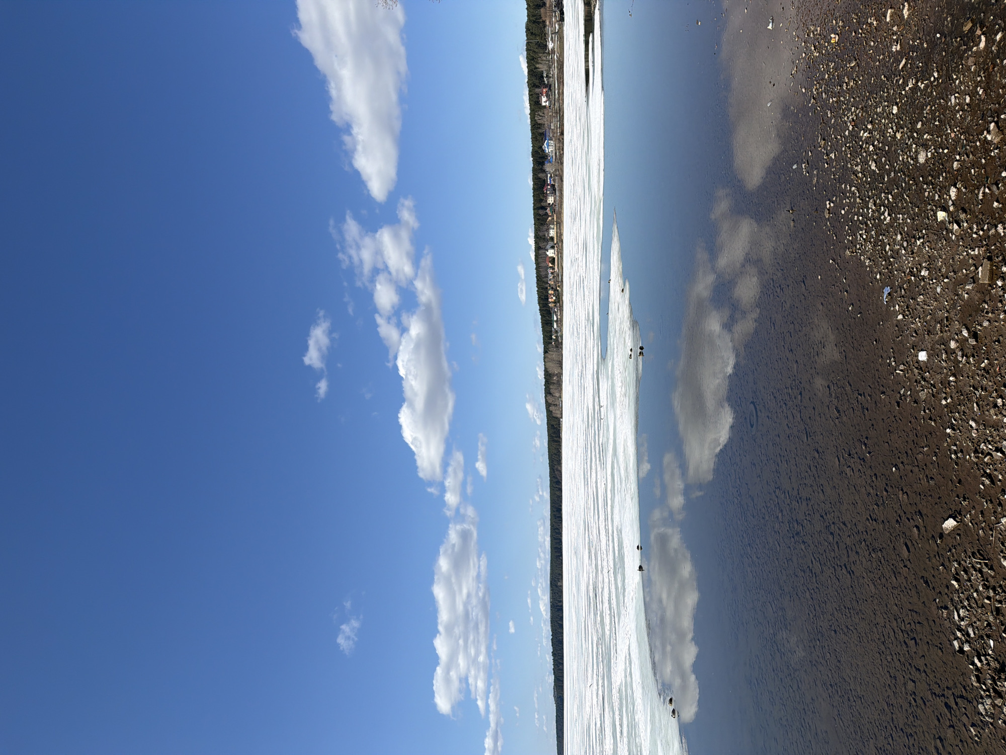 The image depicts a horizontal landscape with a sharp contrast between the natural and man-made elements. On the left, a bright blue sky with scattered white clouds occupies roughly half of the frame. The clouds are varied in shape and size, adding depth to the sky.
The center of the image features a body of water partially covered with ice. The ice extends horizontally across the middle section, reflecting the sky and clouds above. A thin line of trees and buildings is visible in the distance, along the shore of the body of water, suggesting a settlement or urban area.
The right side of the image showcases a shoreline comprised of dark, rocky terrain. The transition from the water to the shoreline is abrupt, emphasizing the difference in texture and color. The water's surface reflects the clouds above, blurring the distinction between the sky and water.
The overall composition suggests a scene captured near a body of water, possibly in a transitional season where ice still lingers. The contrast between the clear sky, the frozen water, and the rocky shoreline creates a visually diverse landscape. It likely represents a location in the city of Votkinsk, Russia, based on the presence of buildings on the horizon, near a river or large lake.