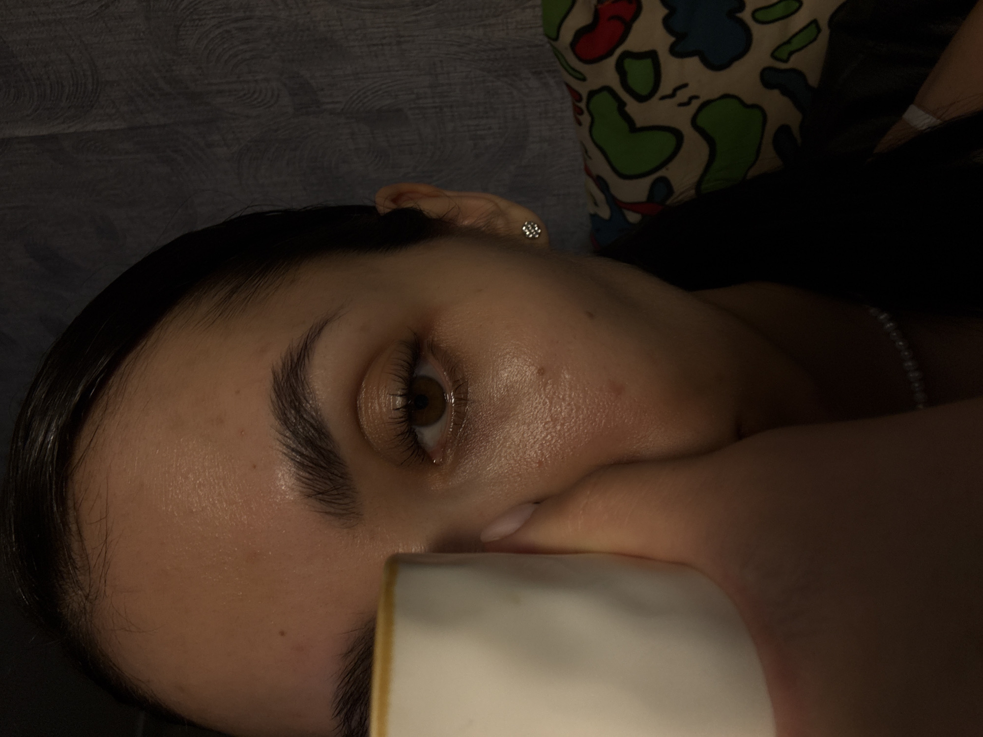 A close-up shot depicts a person lying down, turned towards the camera. Their face is prominent, with one eye visible, featuring a brown iris. The person's hand is partially covering their mouth, and they appear to be holding a white cylindrical object, possibly a mug or cup. A silver earring is visible on their ear. The background consists of a gray textured surface and a patterned object with colors like green, red, white, and blue. The location and city information (Votkinsk, Russia) are not discernible from the image provided.
