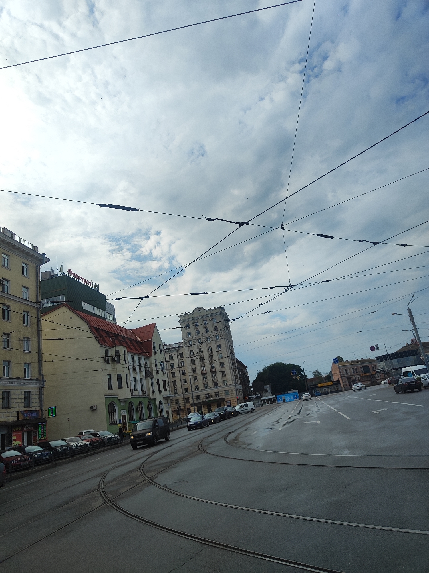 An eye-level, wide-angle shot captures a street scene with trolley wires crisscrossing overhead against a cloudy sky. In the foreground, tram tracks curve to the right, suggesting a junction. The street is wet, reflecting the overcast sky. Several cars are parked along the left side of the road, and a dark minivan is driving on the right lane. Buildings line the street, with the closest on the left being a light yellow, multi-story structure. To its right is a building with a red, pitched roof, and further down the street are more buildings of varying heights and architectural styles, including a tall, ornate building with many windows. A sign on a green building reads "ОКОНТРОЛЬ" in red letters. Traffic lights and street signs are visible in the distance. The overall impression is a somewhat dreary urban environment on a cloudy day.