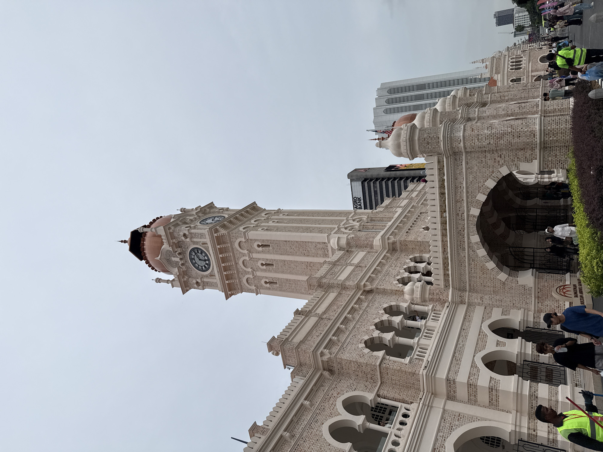 The image depicts Sultan Abdul Samad Building, a landmark building located in Kuala Lumpur, Malaysia. The building's architectural style is predominantly Moorish. It has a clock tower that is visibly displayed. People are observed walking in front of the building. The sky appears overcast. Buildings are visible in the background. A patch of greenery is situated adjacent to the building.