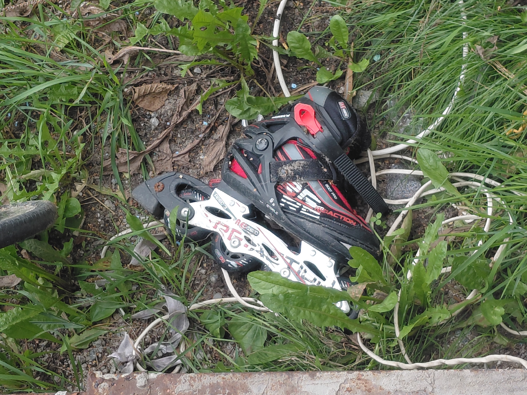 This image depicts a pair of black and white rollerblades lying on the ground amidst grass and dirt. The rollerblades appear worn and have some red accents. A white cable is visible entangled with the rollerblades and the surrounding vegetation. The scene suggests an outdoor environment, possibly a park or a residential area, with a metallic object on the left edge of the frame. The presence of a bicycle wheel in the upper right corner indicates that a bicycle may be nearby. The overall setting is consistent with an urban environment in Nefteyugansk, Russia, though no specific landmarks or identifying features of the city are visible.