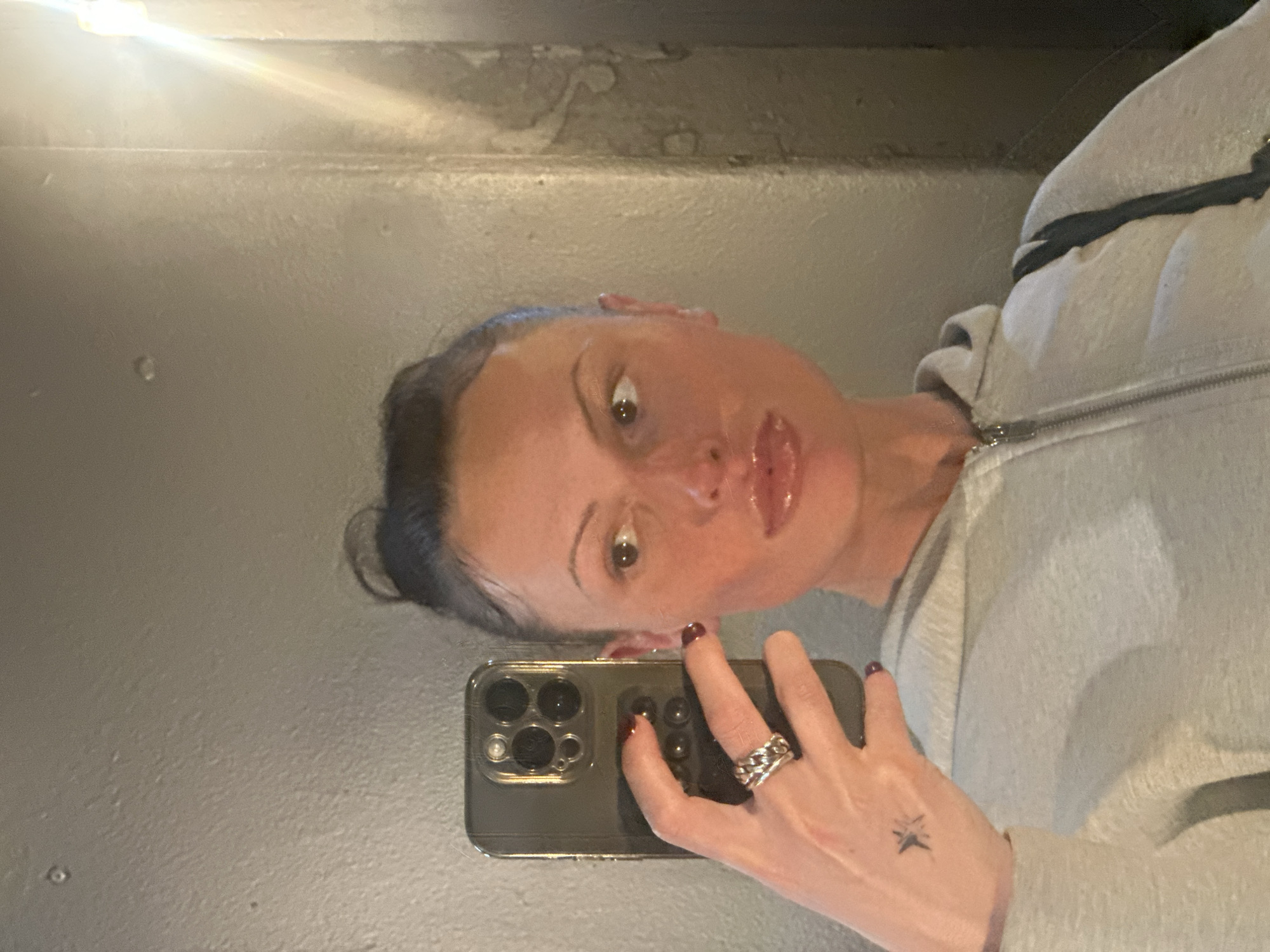 A woman is taking a selfie in what appears to be a bathroom. The woman has fair skin, dark hair pulled back in a bun, and is wearing a gray hooded sweatshirt. She is holding a black iPhone in her left hand, which has a silver braided ring on her ring finger and a small star tattoo on the back of her hand. The background is a gray wall with a textured or slightly weathered surface. There's a bright light source, possibly a light fixture, reflecting off the upper left corner of the wall. The location in the city of London, United Kingdom, cannot be determined.
