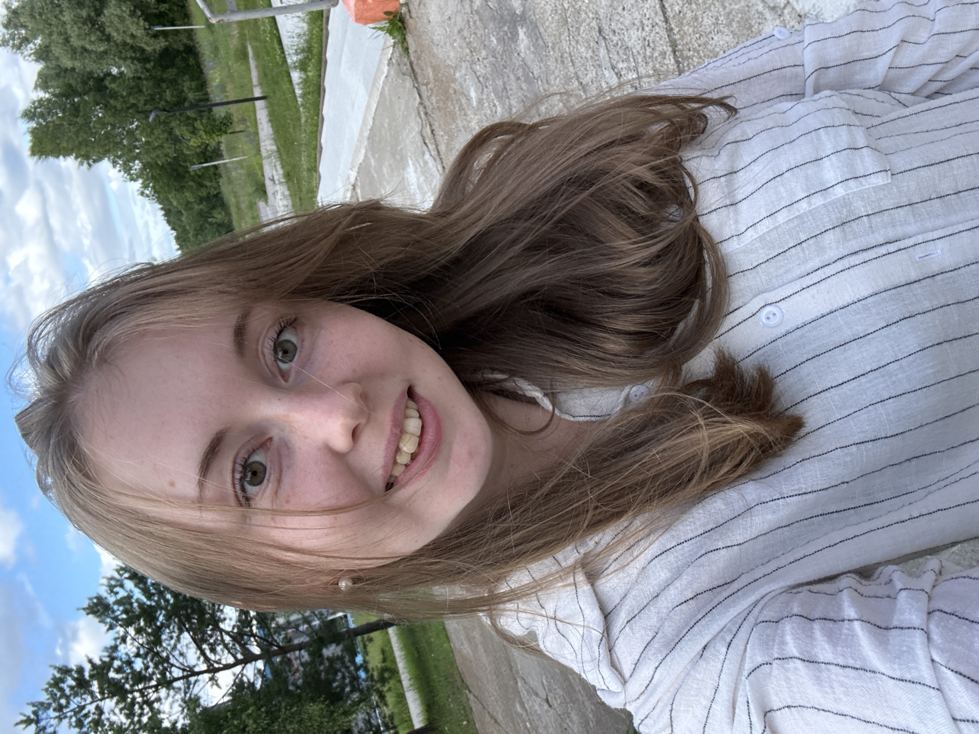 A young Caucasian woman with light brown hair and green eyes is taking a selfie outdoors. She is wearing a white collared shirt with thin black vertical stripes and pearl stud earrings. Her smile reveals slightly yellowed teeth. The background suggests an urban park setting with trees, grassy areas, paved walkways, and a cloudy blue sky. The pavement in the foreground appears worn and textured.