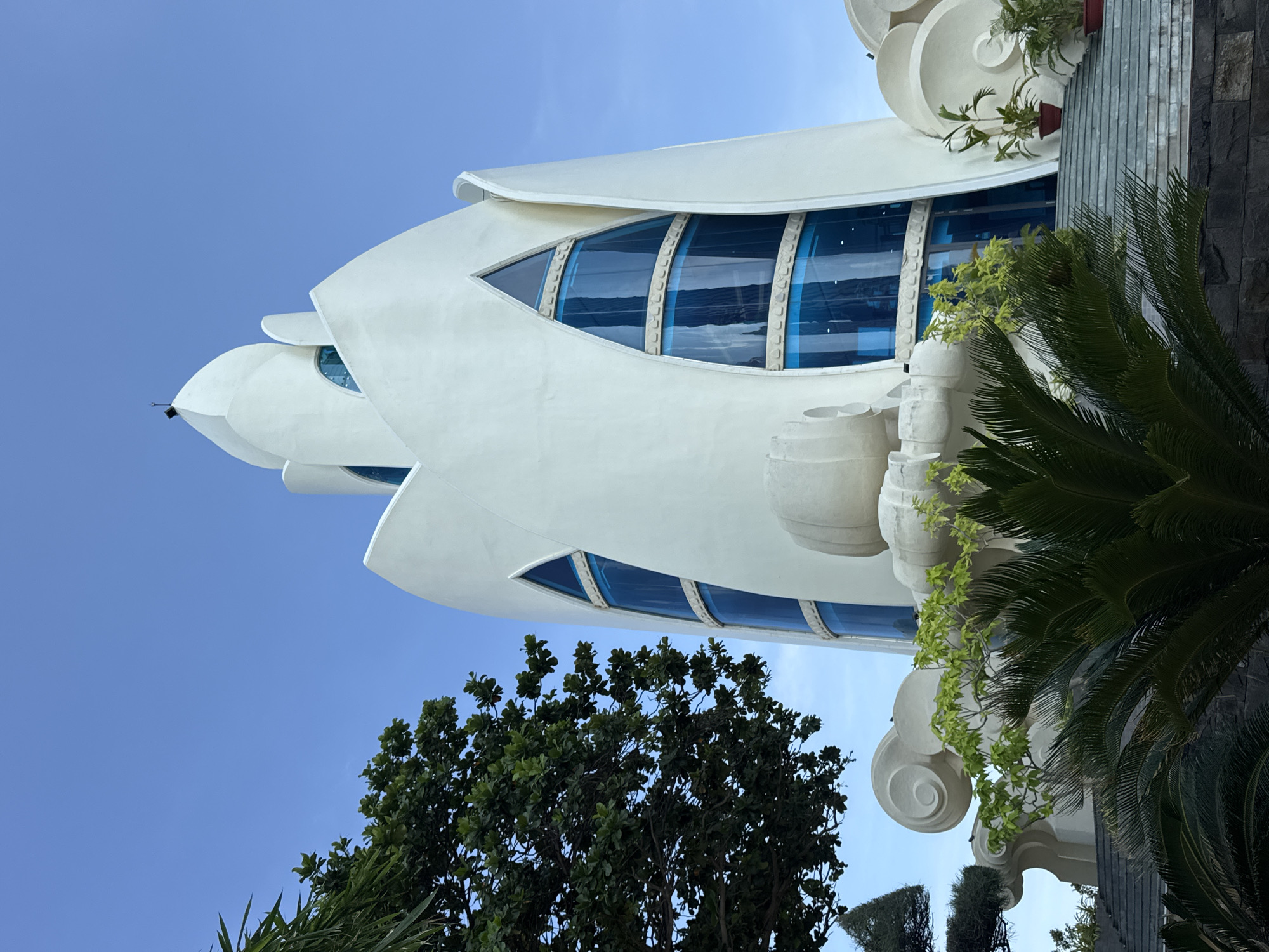 The image shows the exterior of the Long Son Pagoda in Nha Trang Ward, Vietnam. The pagoda is a striking white structure with a tall, tapering tower that resembles a lotus flower. The tower features several large, triangular windows with bright blue glass. The lower part of the pagoda has decorative, cloud-like elements and stairs leading up to the entrance. Lush green trees and plants surround the pagoda, with a clear blue sky overhead. There are no people visible in the image.