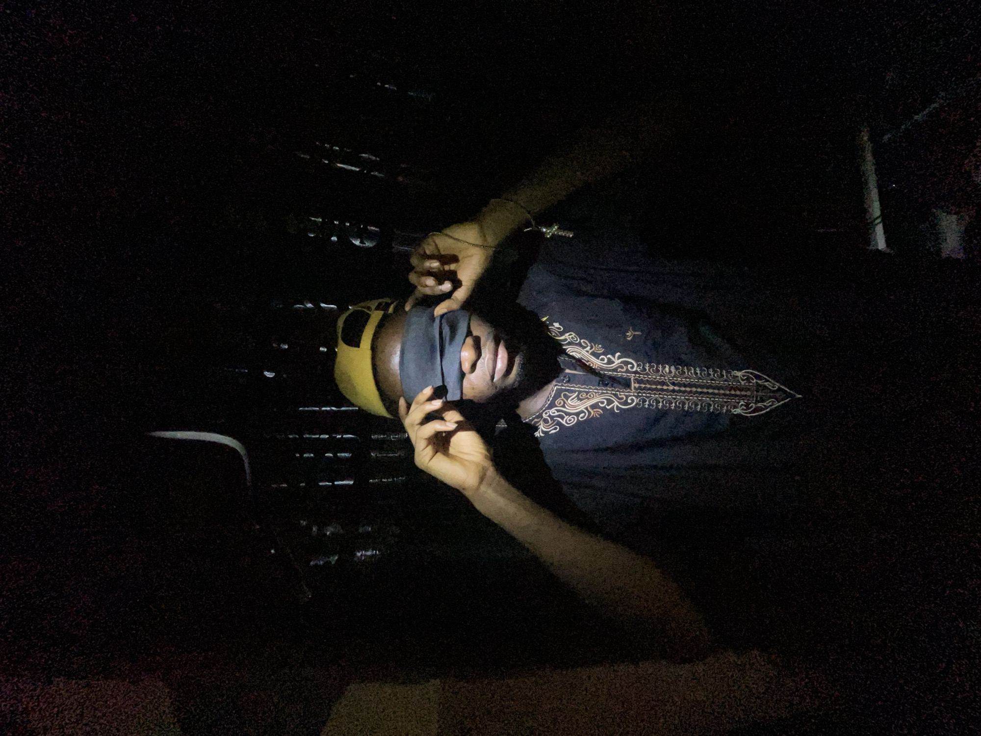 A person with a yellow cap and a black blindfold is lying down in a dark location. The person wears a black shirt with intricate embroidery. Two hands are visible, one holding the blindfold and the other raised slightly. The surroundings are mostly obscured by darkness, with a glimpse of a patterned surface in the background. There is no information present in the image to indicate it is located in Lokoja, Nigeria.