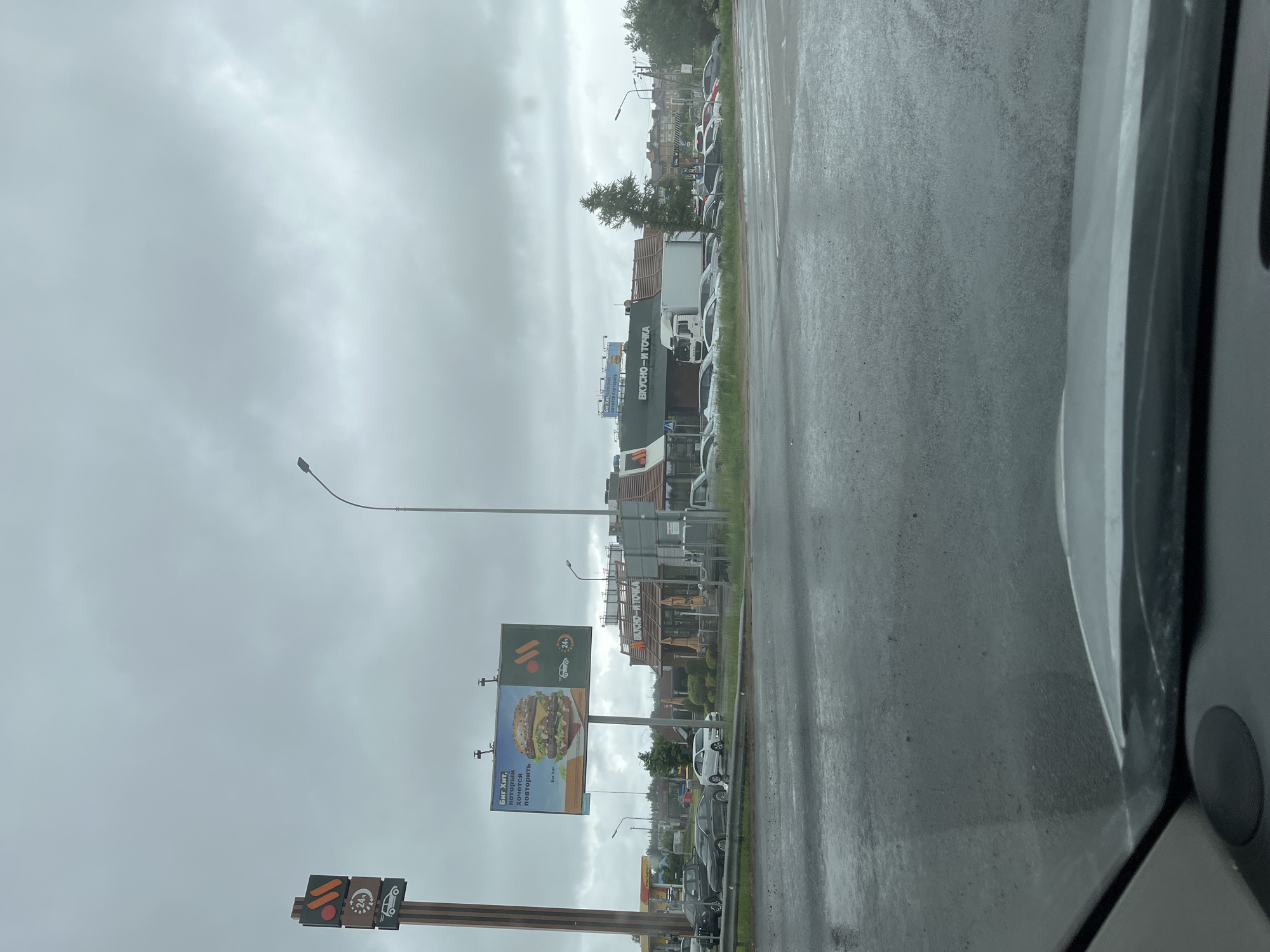 The image depicts a street scene in Мокшино, Russia, captured from inside a vehicle. It is a cloudy day, and the road appears wet, possibly from rain. The primary subject is the "Вкусно - и точка" (Vkusno — i tochka) restaurant, which is the successor to McDonald's in Russia. The building is visible along with a lineup of parked vehicles in its drive-through lane. A large billboard displays a hamburger advertisement, and a traffic sign indicating operational hours and service options is also visible. Other structures and vehicles line the street in the background.