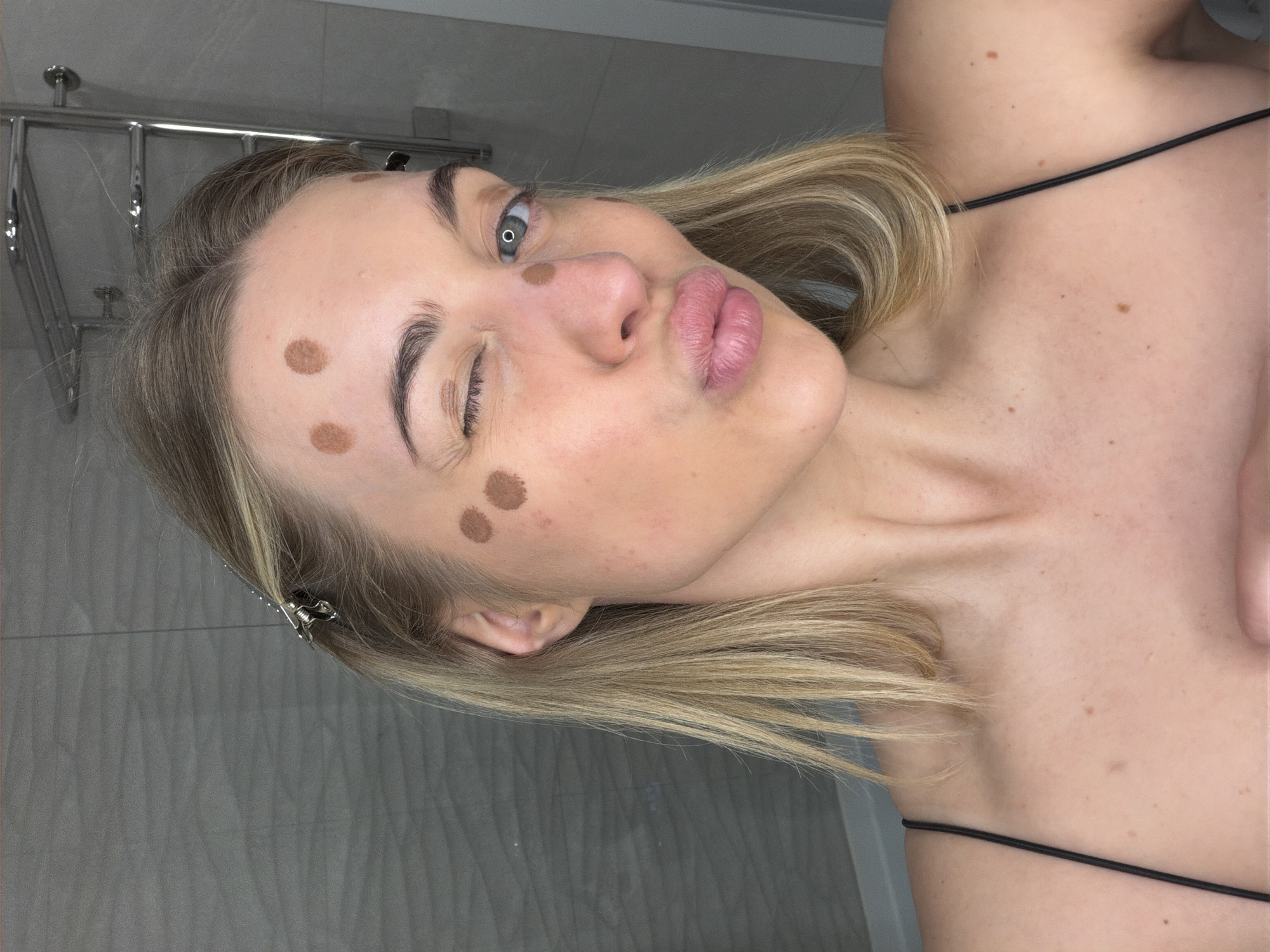 A young woman with blonde hair, light blue eyes, and fair skin is captured in a rotated, close-up shot, likely in a bathroom setting in Sofiivska Borshchahivka, Ukraine. She is seen with several distinct brown dots on her forehead and left cheek, suggesting she is in the process of applying foundation, contour, or a facial spot treatment. Her blonde hair is partially held back by silver clips. The woman strikes a playful pose, winking with her left eye while pursing her lips. She is wearing a top with thin black spaghetti straps, and a few small moles are visible on her neck and chest area. The background features a grey tiled wall and a chrome bathroom fixture, possibly a towel rack. No text is visible in the image.