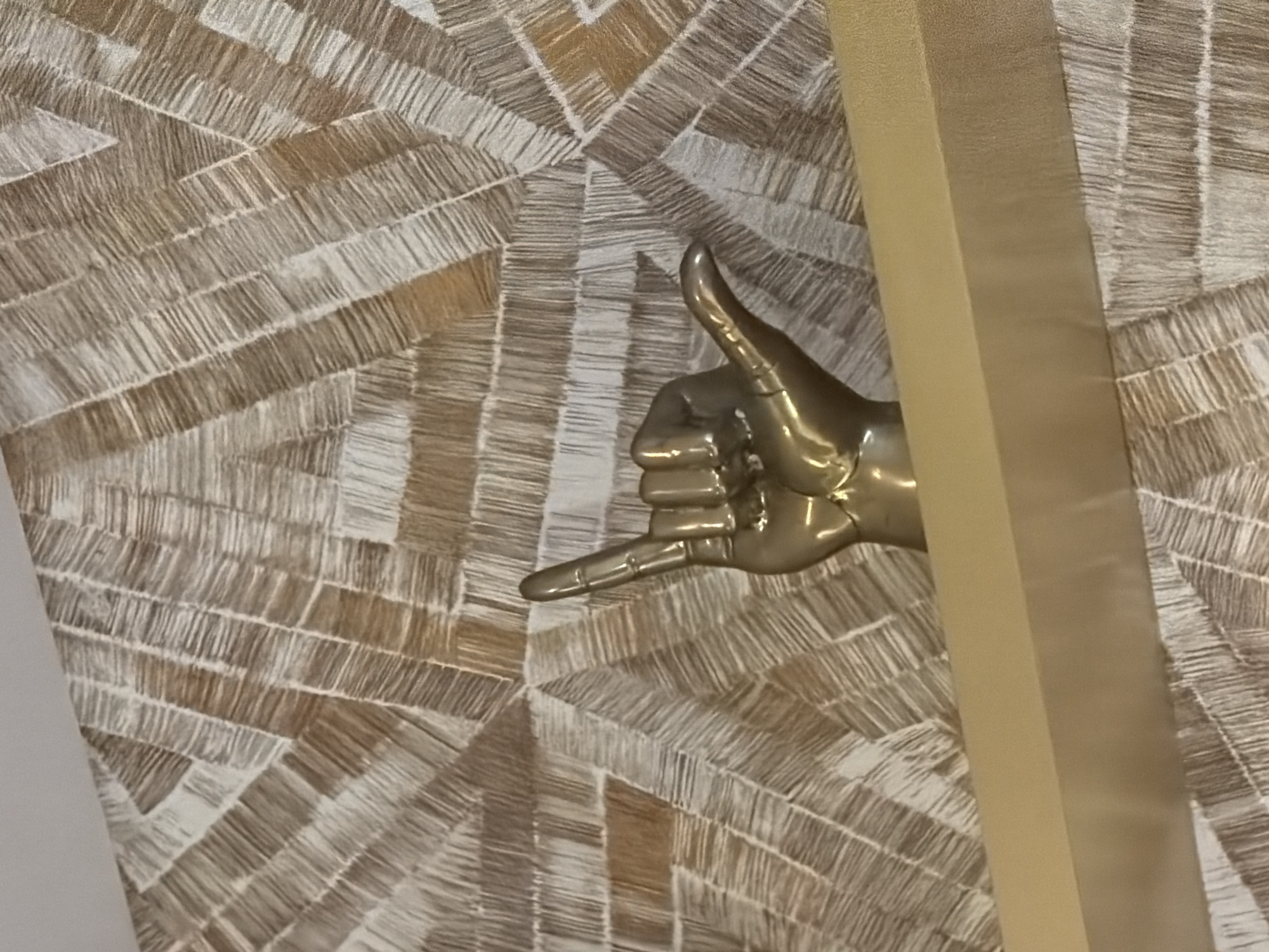 The image features a stylized gold-colored hand making a "shaka" sign, often associated with Hawaiian culture and meaning "hang loose." The hand is positioned in front of a patterned background with geometric shapes in beige and brown tones. A gold rectangular object is partially obscuring the hand. The environment and subjects suggest this could be an interior decor element. Without additional information, a precise location in Kuta, Indonesia cannot be determined.