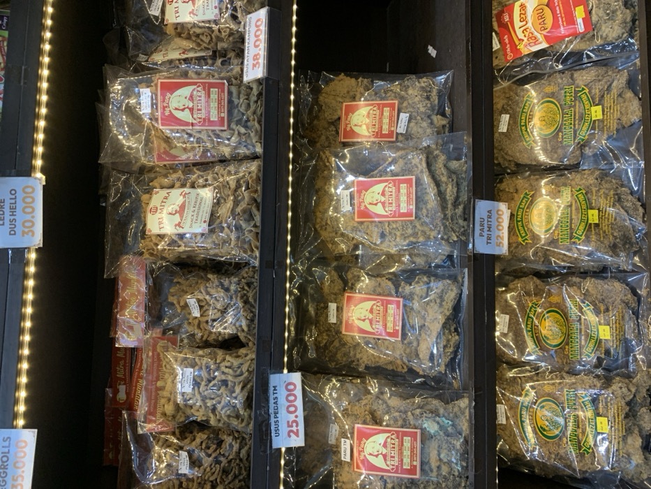 The image shows a display of packaged snacks in a shop located at the coordinates -7.584, 110.287, in Muntilan, Indonesia. There are rows of plastic-wrapped snack foods, displayed on shelves illuminated by bright fluorescent lights. Each package features a brightly colored label with a product name, price, and a small image of the snack. The snacks are all different shapes and sizes, with some resembling chips or crackers, while others have a thicker, more substantial texture. The display is arranged neatly and well-organized, suggesting a tidy and hygienic environment. The pricing is displayed prominently, making it easy for customers to choose their desired snacks.