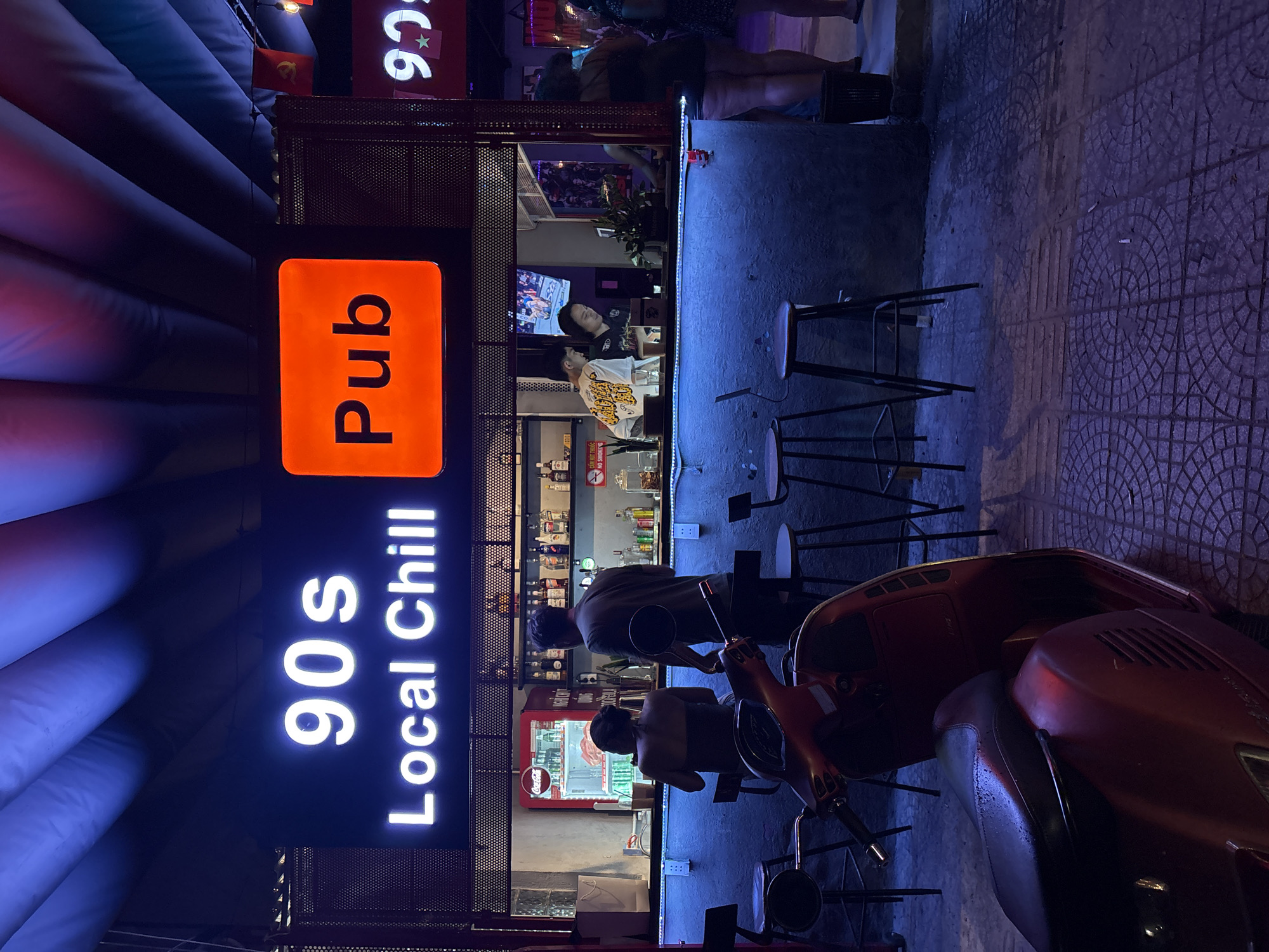 The image depicts an evening scene at a bar named "90s Local Chill Pub," located in Đà Nẵng, Vietnam. The bar's illuminated sign, with an orange "Pub" box, is prominent. Several people are either sitting at the bar or standing nearby. The bar appears well-stocked with bottles and other items on display. A red scooter is parked close to the bar area. The ground is tiled. The environment is dimly lit with a blue hue dominating the scene.