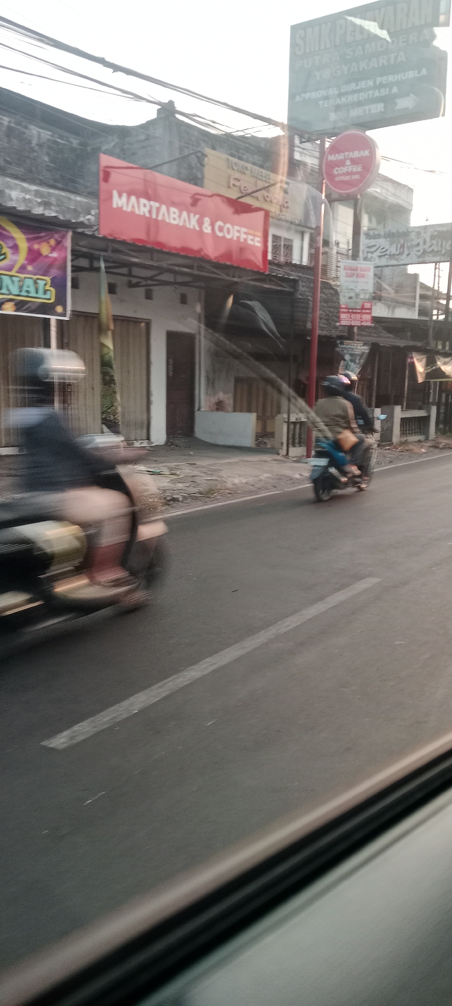 The image captures a street scene in Banyuraden, Indonesia. In the foreground, a person on a moving scooter is blurred due to motion. The scooter is traveling on a paved road with a white line marking. Behind the scooter, several businesses are visible. A prominent red banner reads "MARTABAK & COFFEE". To the right, a green sign displays "SMK PELAYARAN PUTRA SAMODERA YOGYAKARTA" and further text related to accreditation. Another circular red sign with "MARTABAK & COFFEE" is also present. The buildings appear to be shops or commercial establishments lining the street. The environment suggests an urban or semi-urban setting with overhead wires visible. The lighting indicates it may be late afternoon or early morning, with sunlight casting some shadows.