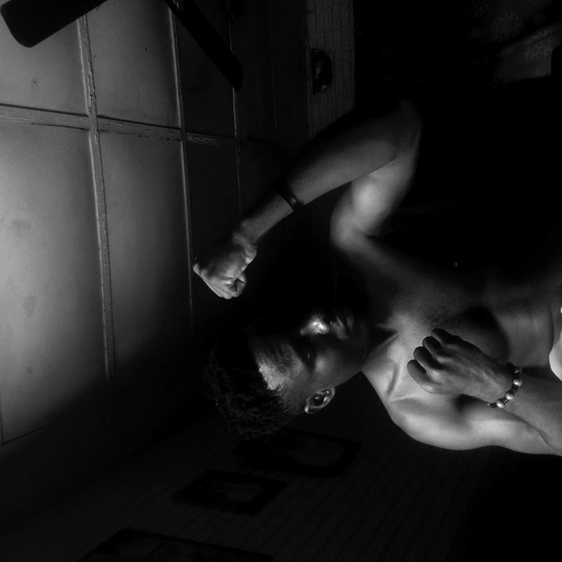 A black and white indoor photograph depicts a shirtless African male from the chest up. His body is angled toward the right, with his face tilted downwards and to the left. He is wearing a bracelet on his right wrist and a wristband on his left. One hand is clenched into a fist, the other is resting on his chest. The background includes a paneled wall to the left and an area of dark indistinct objects near the bottom of the frame. The location is specified as Narayi, Nigeria.
