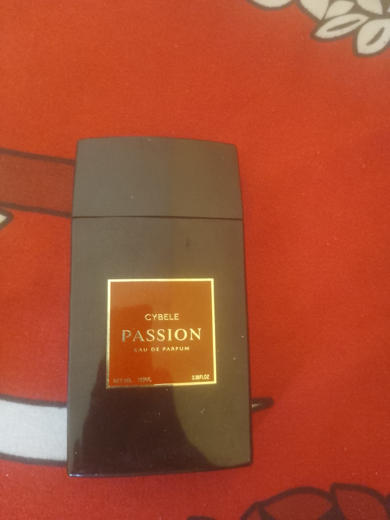 A dark, rectangular bottle of "CYBELE PASSION EAU DE PARFUM" rests on a red surface. The bottle's label is a golden square containing the brand name "CYBELE" in smaller text above the word "PASSION" in large, bold lettering, with "EAU DE PARFUM" below it. At the bottom of the label, "NET VOL 100ML" and "3.38FL.OZ" are visible. The red surface appears to be fabric with a textured pattern. The image does not provide any information or context related to Danbazau, Nigeria.