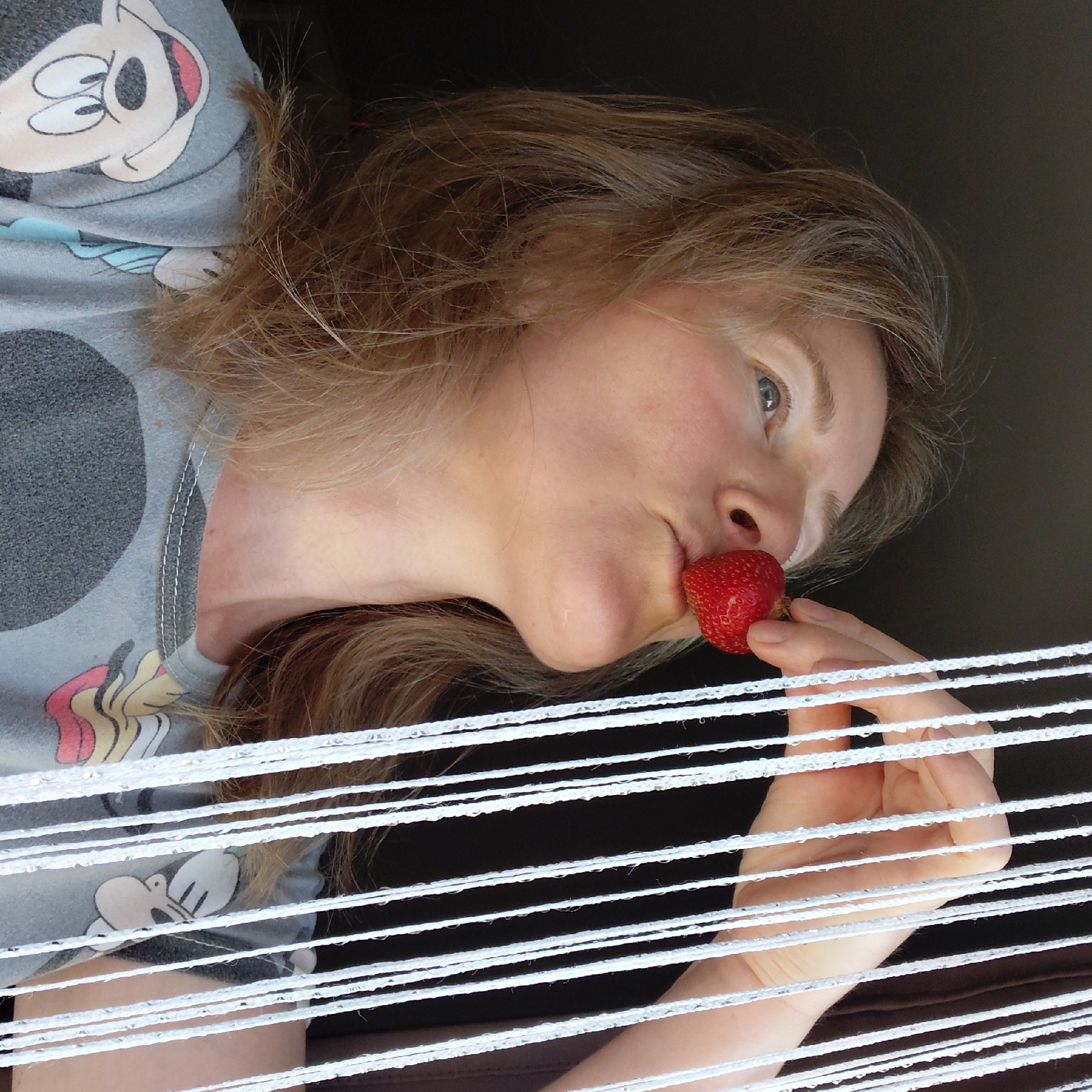 A medium shot shows a woman with blonde hair holding a strawberry to her lips. She is looking upwards and to the left, with her mouth slightly open. Her right hand is visible, holding the strawberry between her thumb and index finger. The woman is wearing a gray t-shirt with a cartoon character print. She is positioned in front of a dark background with white vertical strings hanging down on the right side of the frame. The lighting suggests an indoor environment, possibly near a window. The image does not contain any information to suggest the location is in Novosibirsk, Russia.