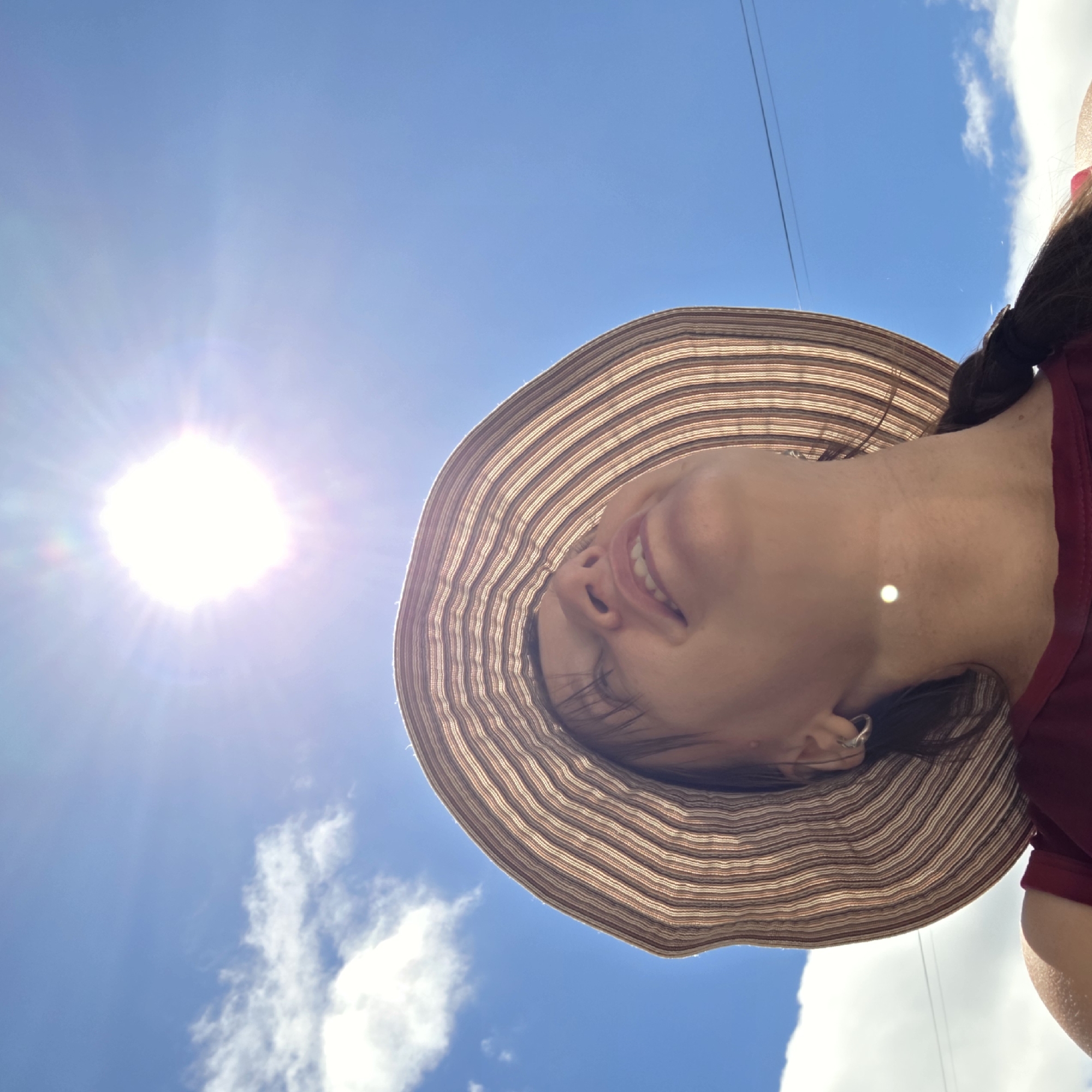 A low-angle shot captures a woman wearing a wide-brimmed, striped hat, looking up at the bright sun in a clear blue sky. Wisps of white clouds are visible. A faint reflection of the sun appears on her neck. She is wearing a maroon top and her dark hair is in a braid. The scene suggests an outdoor setting, potentially in Dunnae-myeon, South Korea, on a sunny day. A thin wire, possibly from a utility line, is visible against the sky in the upper right and mid-right of the frame.