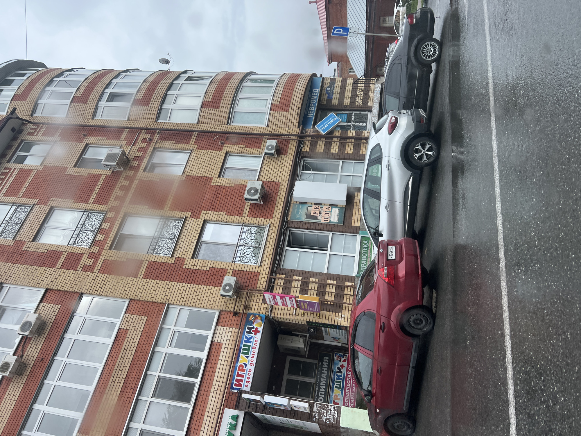 On a rainy day in Ulyanovsk, Russia, a three-story brick building with a curved facade faces a wet asphalt road. The building's exterior features a red and light brown brick pattern, with multiple windows and air conditioning units visible. Several shops are located on the ground floor, indicated by signs such as "ИГРУШКИ" (Toys) and "НЕВРОМЕД" (Neuromed). A medical center sign with the address "ул. Баумана, 10" (Bauman St, 10) is also present. Three cars are parked along the road: a red Chevrolet Aveo on the left, a silver Renault Captur in the middle, and a black Chrysler Sebring on the right. The road surface is reflective due to the rain, and a white line marking a lane divider is visible. In the background, a red-tiled roof of another building and a blue parking sign are visible.