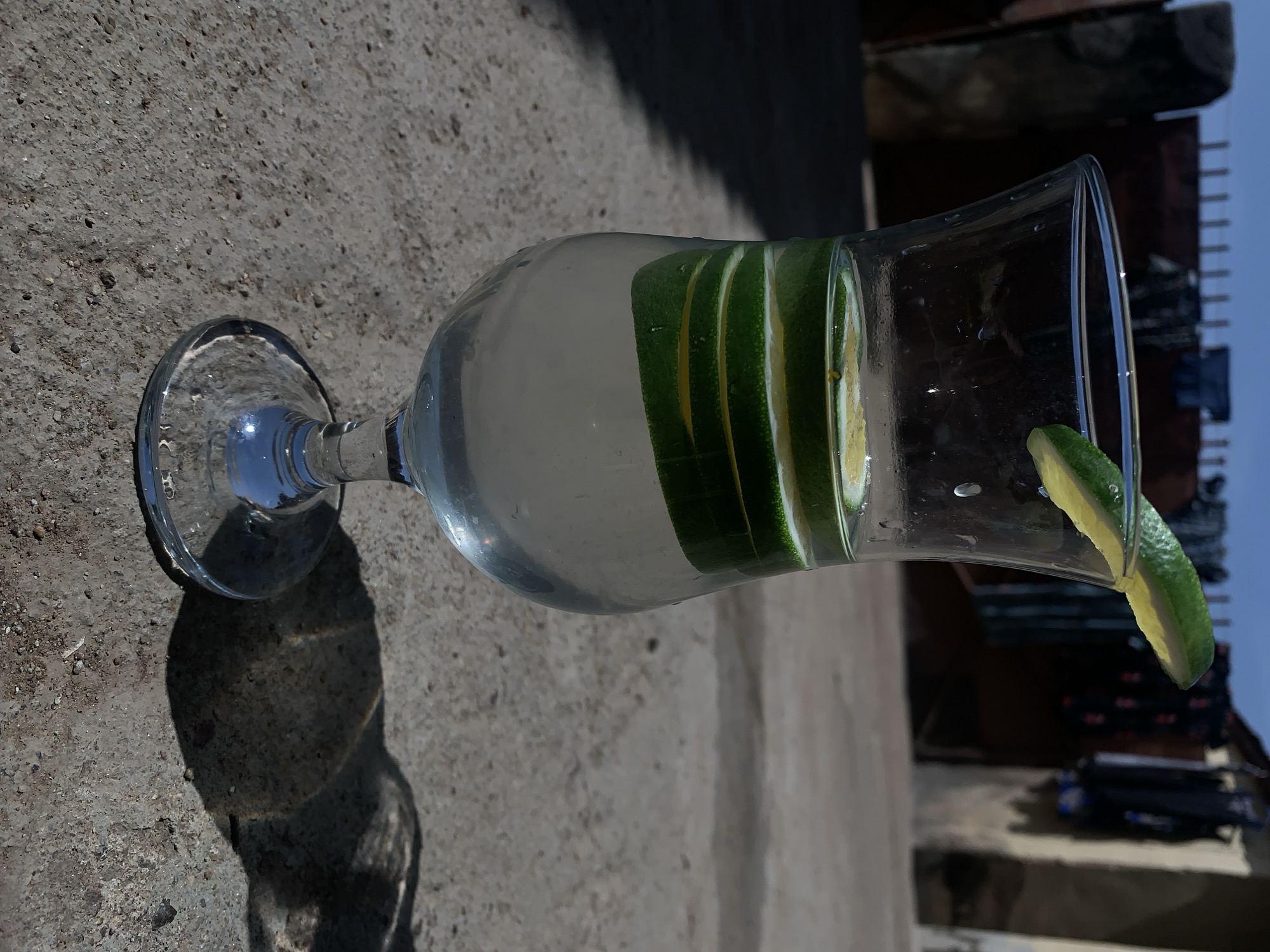 A glass of water with lime slices is resting on a concrete surface outdoors. The glass is tall and slender, with a clear liquid and several lime slices inside. A lime wedge sits on the glass rim. The concrete surface is textured and appears weathered. The background features a building or structure, partially obscured by shadow. The sky is visible in the upper right corner. The location is outdoors in Rafin Par, Nigeria.