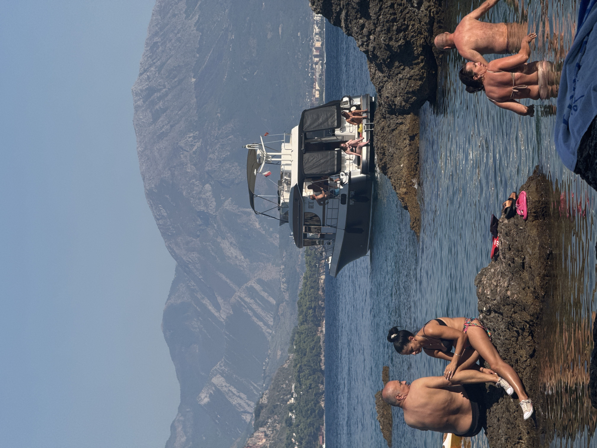The image depicts a sunny day at the beach in Sutomore, Montenegro. In the foreground, two people are seated on rocks in the shallow water, a man on the left and a woman on the right. The man is bare-chested and wearing black shorts, while the woman is in a bikini. Further to the right, another couple stands in the water, a man with his back to the camera and a woman facing away from it. In the mid-ground, a boat is anchored offshore, with several people visible on its deck. The background features a picturesque coastline with lush green vegetation and buildings, leading up to a mountain range under a clear blue sky. The water is calm and reflects the sunlight, creating a serene atmosphere.