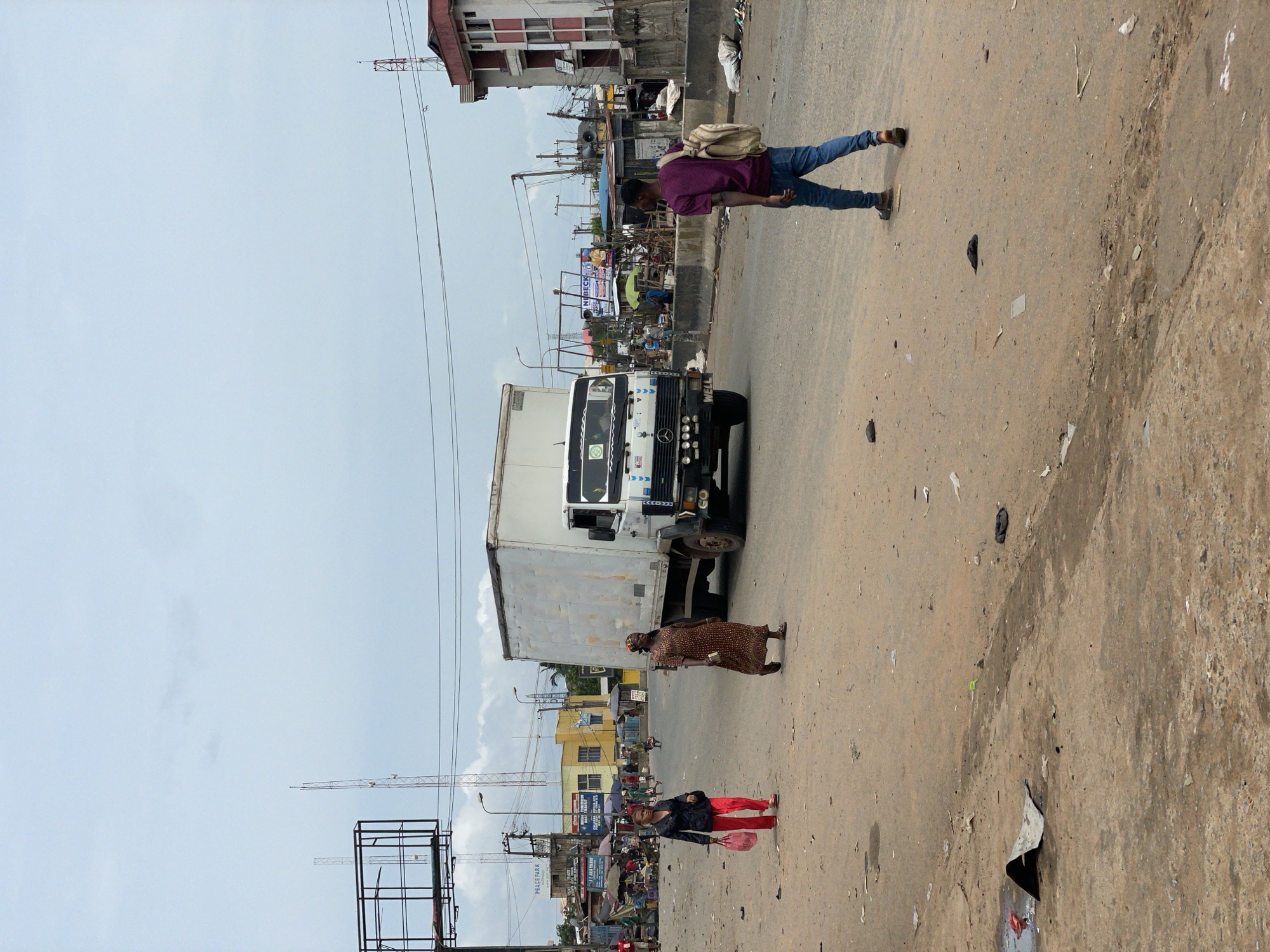 The image depicts a street scene in Nkpor, Nigeria. There are three people visible: a man wearing a purple shirt and jeans, a woman in a brown patterned dress and head covering, and another woman with red pants and a dark jacket. They are walking on a dirt road. A white truck is parked on the road. In the background, there are buildings, signs, market stalls, and overhead power lines. The sky is light blue with some cloud cover. There's construction equipment (crane) in the background. The road surface shows signs of wear and disrepair.