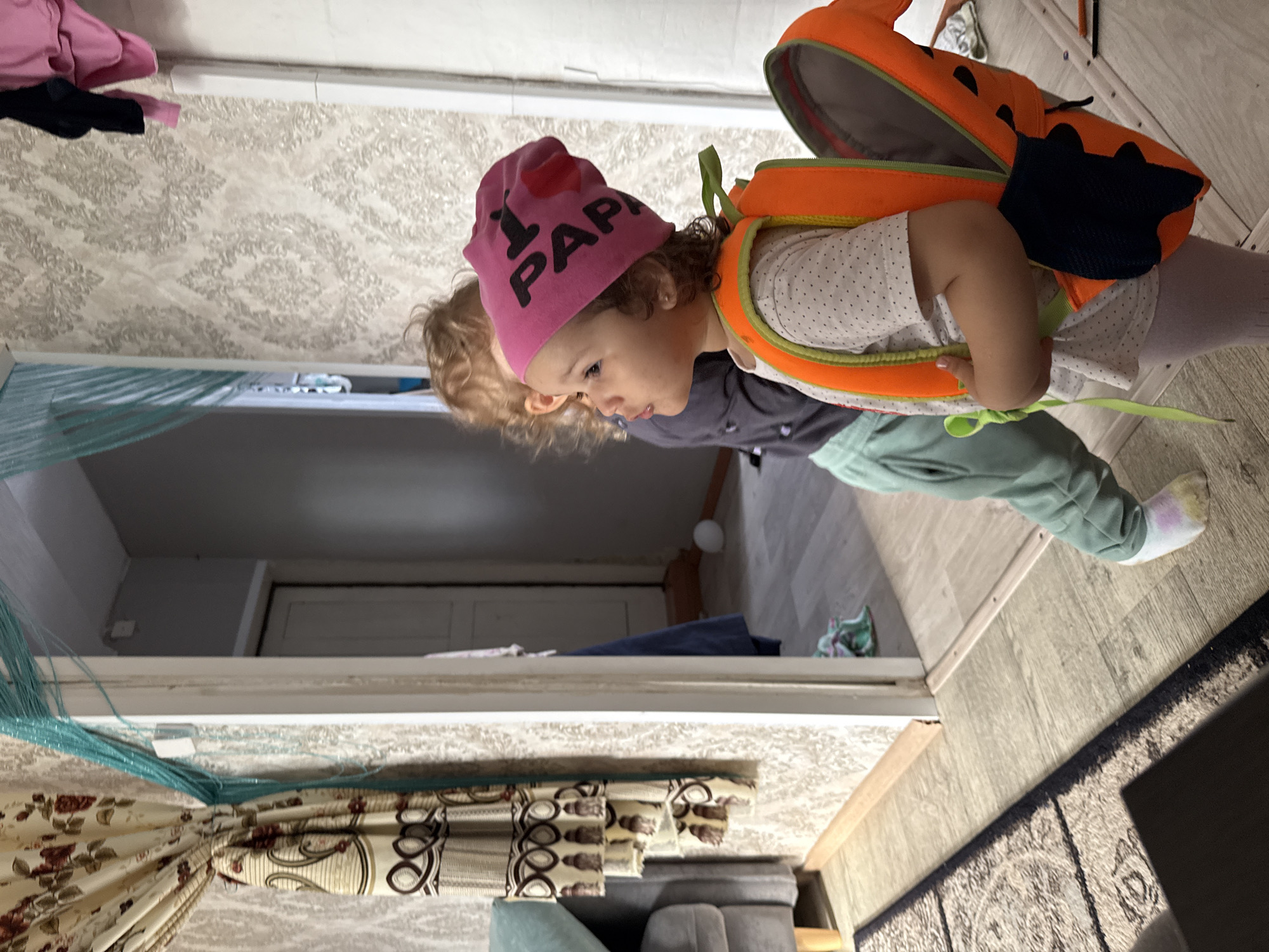 The image, originally rotated 90 degrees counter-clockwise, captures a candid indoor scene featuring a young child, likely a toddler, as the main subject. The child has light brown curly hair and wears a vibrant pink beanie adorned with a red heart symbol followed by the text "PAPA," indicating "I love Papa." They are dressed in a white short-sleeved shirt with small black polka dots and light green sweatpants. An eye-catching orange backpack, possibly designed to resemble an animal with black spots or features, is strapped to their back with lime green accents.
The child is depicted standing, looking downwards with a focused or contemplative expression. Partially visible behind them is another child with lighter hair, also looking downwards. The setting appears to be a domestic interior, possibly a living room or hallway in a home located in Уральское, Russia.
The background features light-colored wallpaper with a subtle, textured pattern. The floor is made of light wooden planks, partially covered by a dark, patterned rug. To the left, patterned curtains with floral and geometric designs are visible, accompanied by light blue or green decorative strings hanging nearby. A piece of grey furniture, possibly a sofa or armchair, is at the bottom of the frame. In the distance, a doorway leads into another room with a darker floor and a white door. The lighting suggests daytime indoors, contributing to a casual and relaxed atmosphere.