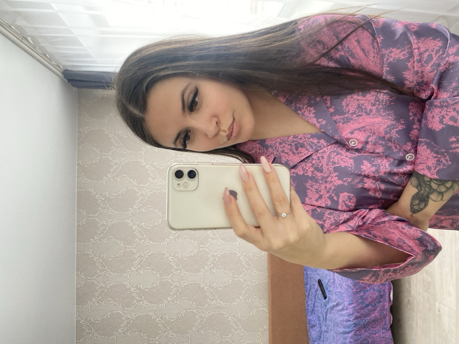 The scene depicts an interior room, identified as being within the city of Chaikovsky, Russia. A young adult female is centered, holding a light-colored smartphone in her left hand to capture a mirror selfie. She has long, dark hair parted in the middle, dark prominent eyelashes, and her lips are slightly pursed. She is dressed in a collared top featuring a purple and pink floral or toile de Jouy pattern with light-colored buttons. Her left hand exhibits long, light pink manicured nails and a ring with a small, light-colored stone on her ring finger. A portion of a dark, coiled tattoo is visible on her inner right forearm, above the sleeve cuff. The background, reflected behind the subject, features a wall covered in light-colored wallpaper with a subtle, ornate damask pattern. To the left, a section of a brown headboard or sofa back is visible, beneath which a bed or sofa is covered with a purple and blue patterned blanket. A dark remote control rests on this blanket. To the right, a window with sheer white curtains and a darker blue-grey curtain allows natural light into the room.