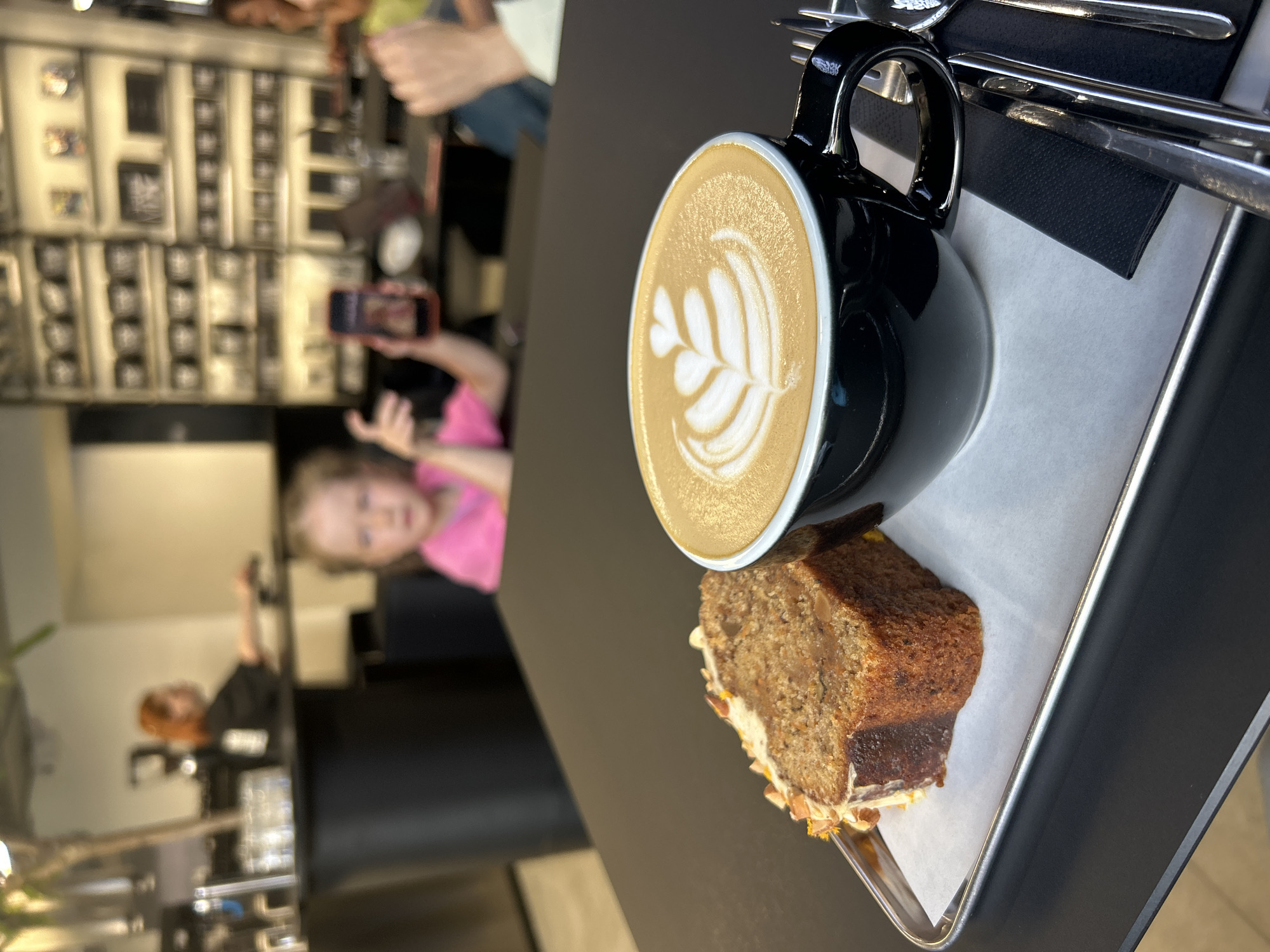 The image shows a close-up of a black mug with latte art, filled with a coffee drink, placed on a metal tray next to a slice of cake. The mug and cake are positioned on a dark table, with a blurred background. In the background, there are several people, including a young girl in a pink shirt who is looking at a phone, and another person with red hair. The scene appears to be in a cafe or restaurant. The lighting suggests it could be daytime. The overall mood is casual. There is no visible text in the image. The location context provided is Izhevsk, Russia.