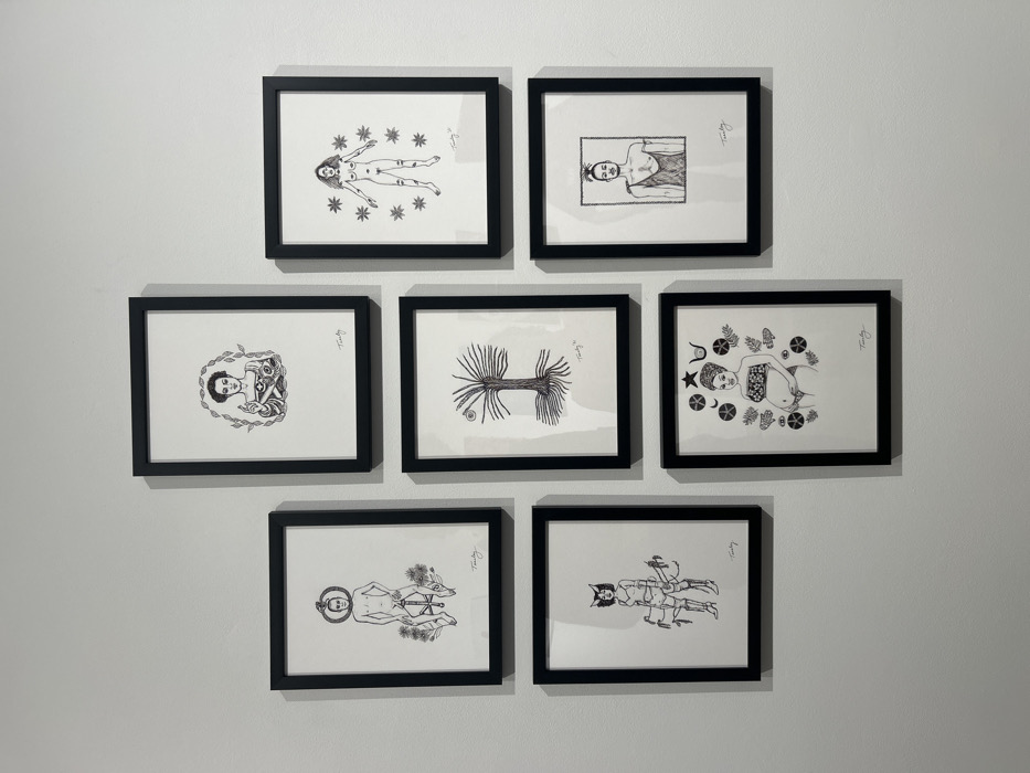Seven framed black-and-white ink drawings are displayed on a plain white wall, likely within a gallery or exhibition space in the city of Los Angeles, United States of America. The frames are black, each enclosing a white matte around the artwork. The arrangement consists of one central drawing at the top, three drawings in the middle row, and three drawings in the bottom row, forming a vertical staggered grid.
Each drawing is a stylized illustration, typically depicting human or human-like figures or symbolic elements. All visible artworks bear the signature "Tunty" in the bottom right corner.
**Artwork Details (from top to bottom, left to right):**
1. **Top Center:** A head-and-shoulders portrait of a woman with dark, wavy hair encircled by a laurel wreath. She wears a patterned top and holds a small, spherical object between her hands.
2. **Middle Row - Left:** A full-body depiction of a nude, slender figure, possibly male, with a serpentine or ring-like object encircling its head. The figure stands atop a circular base amidst stylized flowers and foliage.
3. **Middle Row - Center:** A drawing of a stylized tree with extensive, exposed roots. An oval shape, possibly an eye or fruit, is depicted at the apex of its branches.
4. **Middle Row - Right:** A full-body depiction of a nude, long-haired figure, possibly female, marked with lines on the thighs and calves. The figure is surrounded by twelve six-pointed stars.
5. **Bottom Row - Left:** A full-body depiction of a bipedal figure, possibly female, with pointed, animal-like ears and an elaborate, armored costume including gauntlets, knee-high boots, and a chest plate. The figure holds two long, thin objects resembling spears or staffs.
6. **Bottom Row - Center:** A semi-nude depiction of a pregnant woman with styled hair. She wears a patterned bandeau top and is surrounded by various symbolic elements including a crescent moon, a star, a bull's head, and botanical motifs.
7. **Bottom Row - Right:** A bust portrait of a figure, possibly male, with a top knot hairstyle and facial hair. The figure wears a garment draped over one shoulder, exposing the chest. The background behind the figure features a dense, textured pattern.