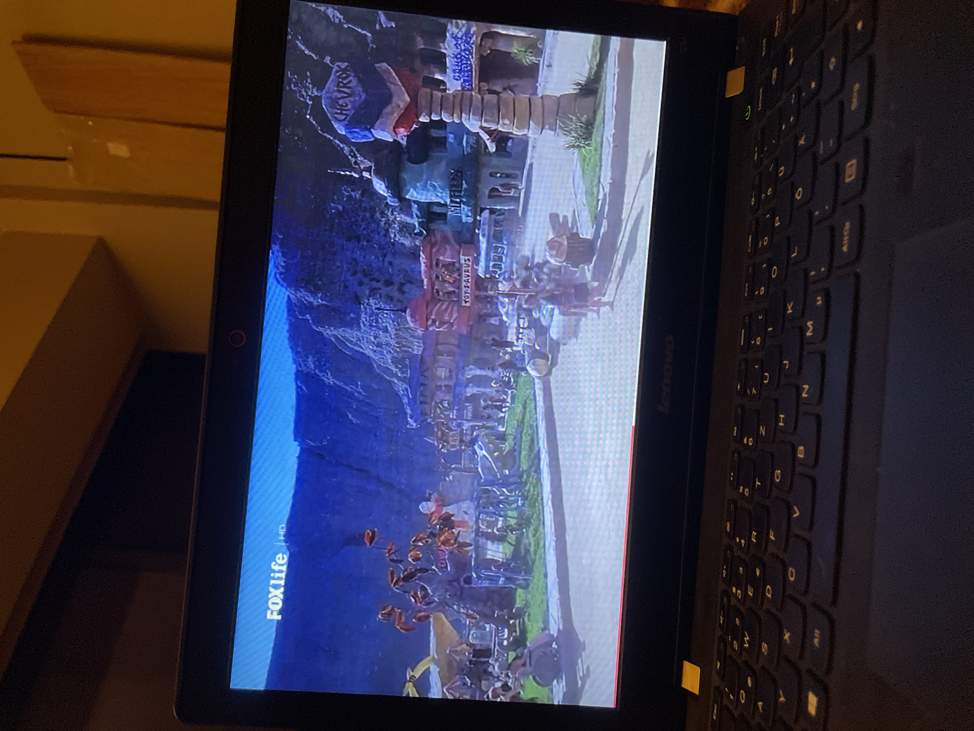 Here is a description of the image:
The image shows a laptop displaying a scene seemingly set in Πόρτες, Greece. The display features a mountain range. In the foreground, there are signs with writing including "YOT-S-AVRUS" and "CHEVROK". There appear to be many people in the streets. The image is being displayed on a Lenovo laptop. The screen also has the Foxlife HD logo in the corner.