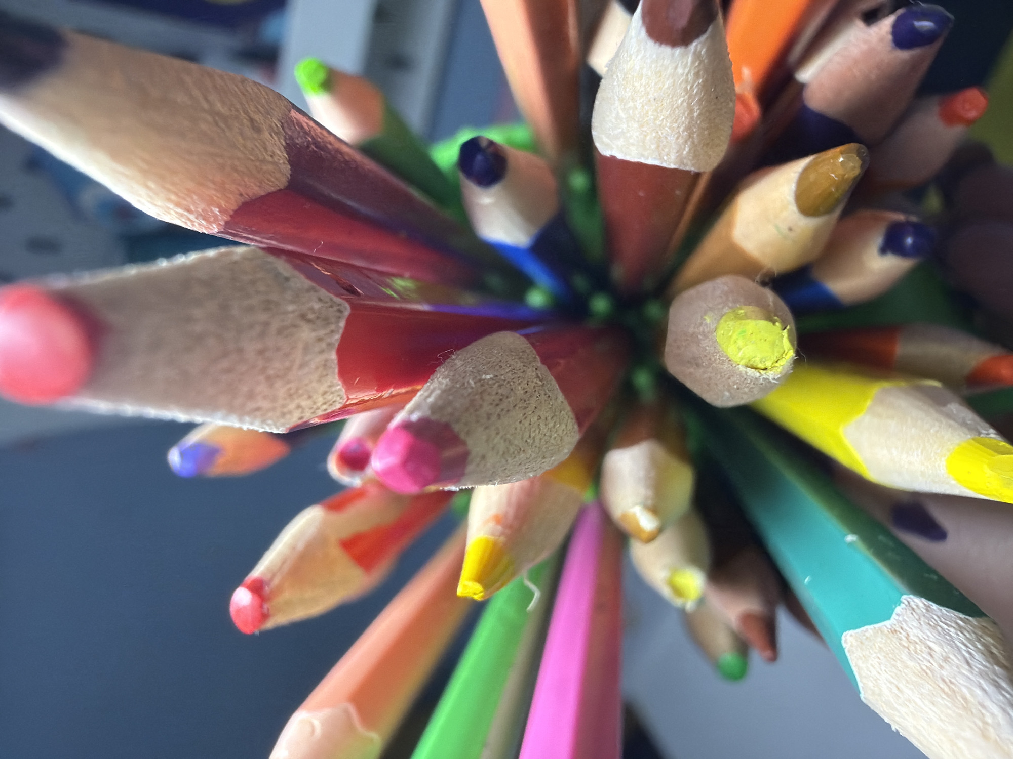 The image is a close-up, macro shot of the sharpened tips of a collection of colored pencils. The pencils are arranged in a bouquet-like fashion, with their points directed upwards and outwards. Various colors are visible, including red, pink, yellow, green, blue, orange, and purple. The wood grain of the pencils is textured and detailed, and the colored lead tips are sharp and vibrant. The background is a dark, blurry blue, suggesting an indoor setting, possibly a desk or art supply area. There is no visible connection to Votkinsk, Russia, as the image is a generic still life of art supplies.