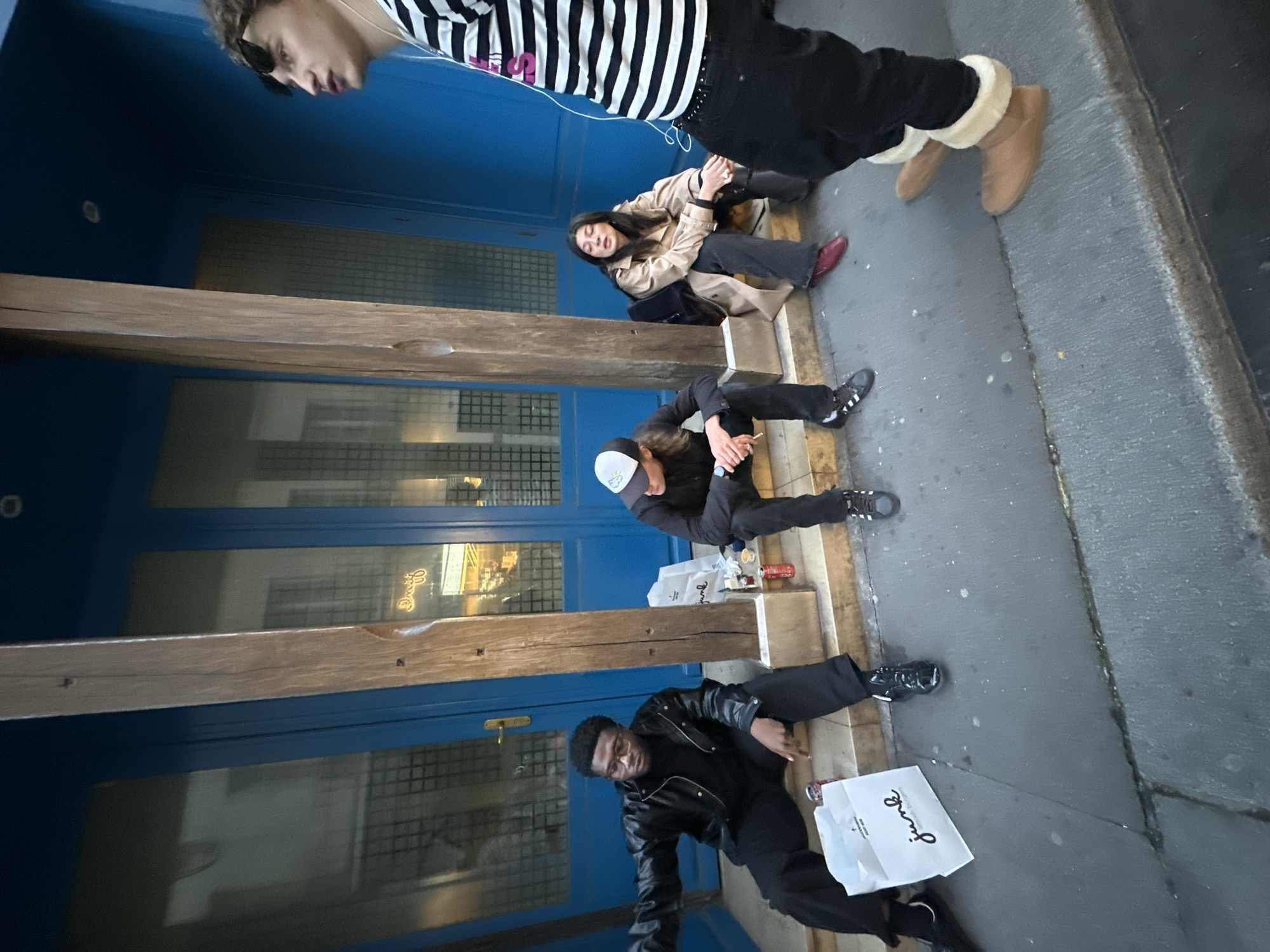 The image depicts a group of four people sitting or lying on the sidewalk in front of a building. The building has a blue facade with windows, and wooden beams. The people are dressed in casual clothing, including jackets, pants, and boots. One person is wearing a striped shirt and sunglasses. Another is holding a white shopping bag that reads "Siuiz". There are also some beverage containers visible on the sidewalk. The location appears to be a street in Paris, France, given the architectural style.