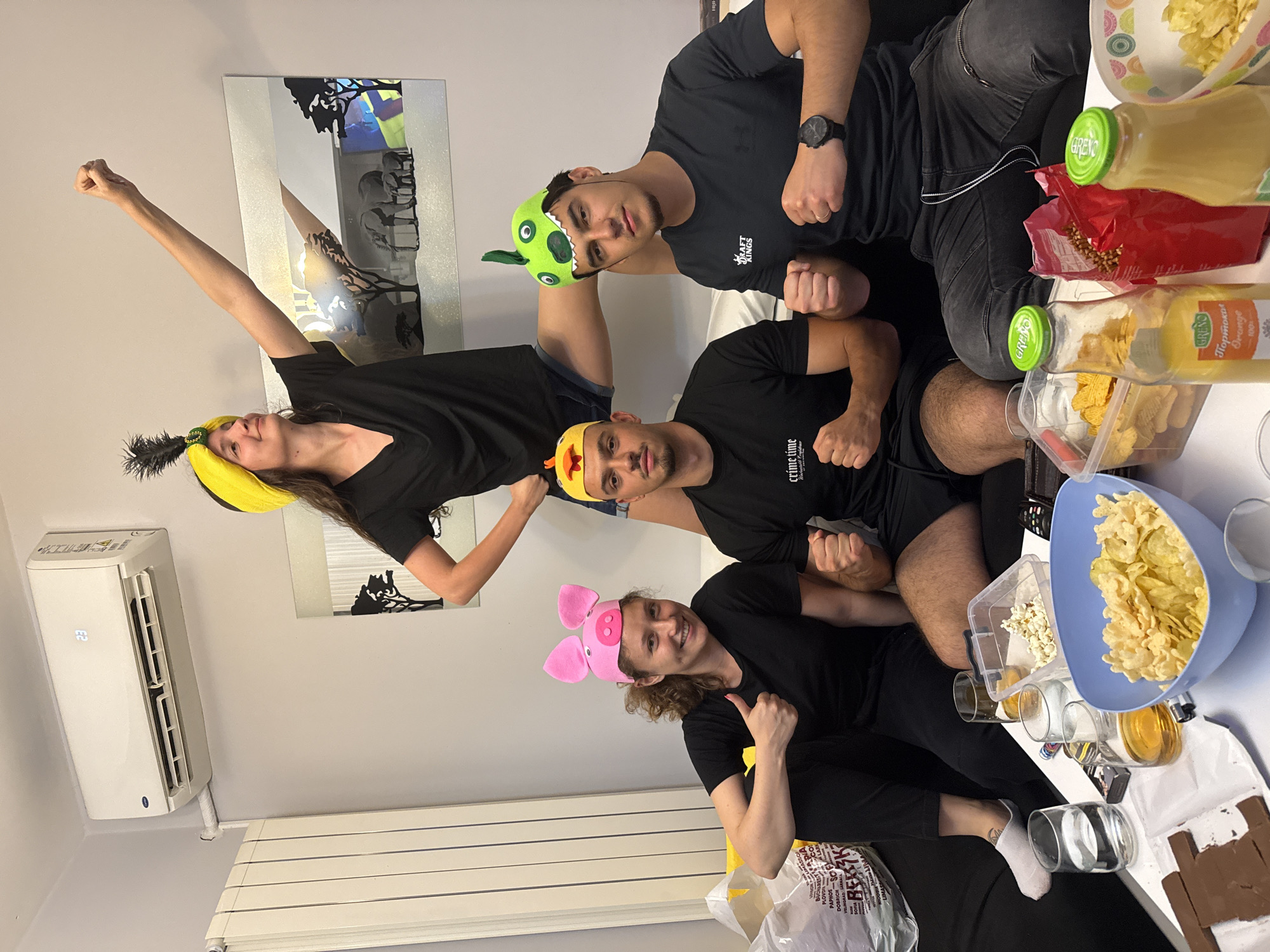 Four people are captured in a vertical-oriented indoor shot, appearing to be at a casual gathering or party. Three men and one woman are sitting closely together, all wearing black t-shirts and novelty hats. The person on the far left wears a yellow and black feathered hat, with their arm raised high and fist clenched in a celebratory gesture. Next to them, a man wears a green monster-like hat. Below him, another man sports a yellow chicken hat, while the woman on the right wears a pink pig hat and gives a thumbs-up. Most individuals have clenched fists, adopting playful, strong poses and smiling towards the camera.
The scene includes a variety of snacks and drinks placed on a table in front of the subjects. These include a blue bowl filled with potato chips or crisps, a clear container of popcorn, and two bottles of orange juice labeled "GRENO Портокал Orange 100%". Several glasses are also present.
In the background, a light-colored wall features a decorative reflective piece with dark, tree-like silhouettes. An air conditioner unit showing the number "32" is mounted on the wall, with a white radiator visible below it. Visible text on clothing includes "crime time" and "DRAFT KINGS". A white plastic bag on the floor is marked with "BERK" and a list of city names: "PLOVDIV", "PAPHOS", "DOBRICH", "VELINGRAD", "LARNACA", "LIMASSOL", and "SOFIA", indicating the location context of Sofia, Bulgaria. The overall atmosphere is joyful and relaxed.