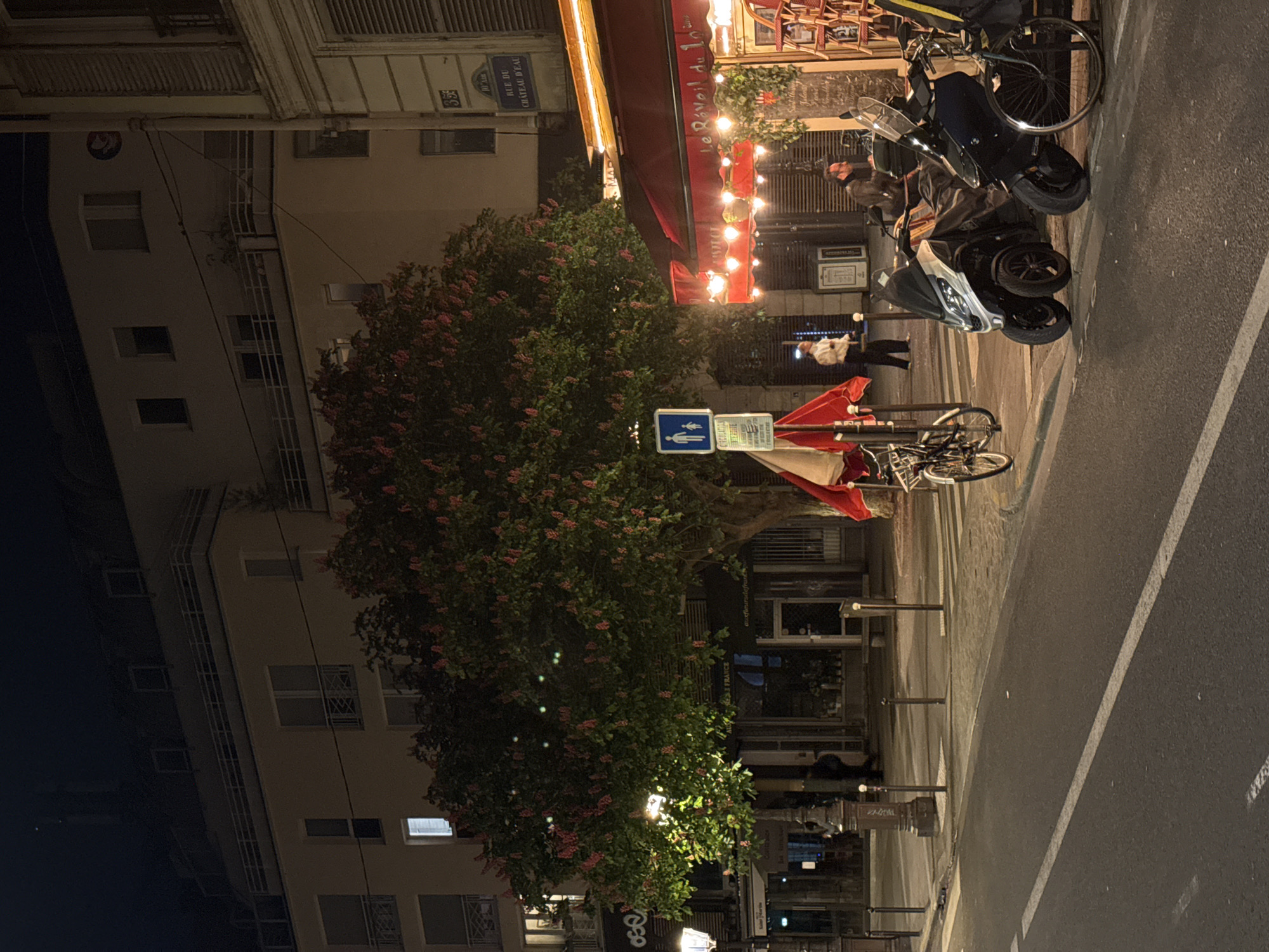 The image depicts a street scene at night in Paris, France. On the right side of the frame, there is a street with parked motorcycles and bicycles. A red awning covers an establishment called "Le Réveil du 10ème," adorned with strings of lights. People can be seen near the establishment. On the left, there is a tall building with multiple windows, some illuminated. A large tree with blooming flowers is centrally positioned, partially obscuring the building and street. Street signs are visible, along with various street furniture such as lamp posts and traffic signs.