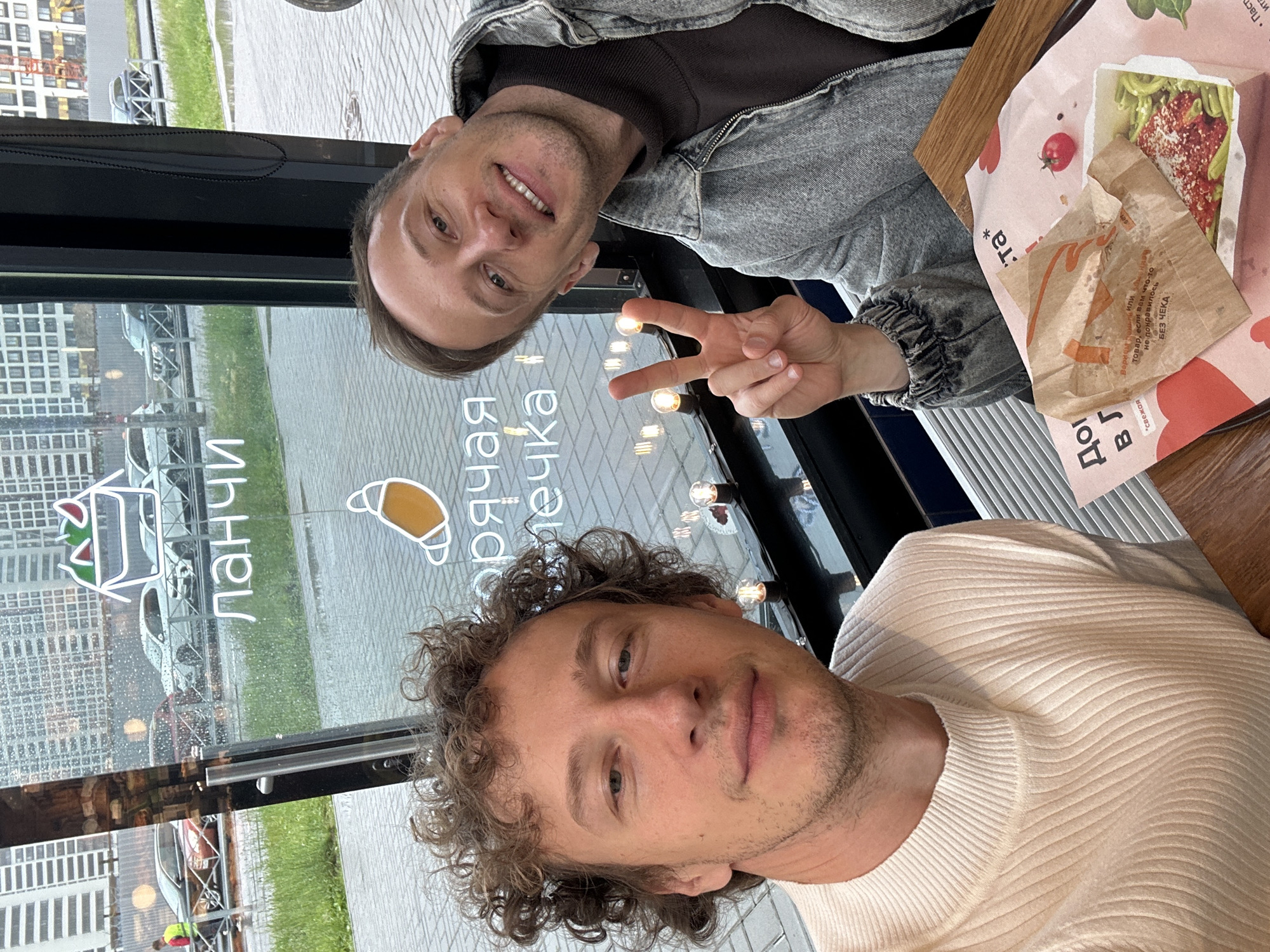 Two men are shown in a selfie taken from a low angle, with one man in the foreground and the other slightly behind him. The man in the foreground has curly brown hair and is wearing a cream-colored ribbed sweater. He is smiling faintly. The man behind him has short brown hair and is wearing a denim jacket over a dark shirt. He is smiling broadly and making a peace sign with his fingers.
They are seated at a table, possibly in a cafe or restaurant, as suggested by the presence of food items and wrappers on the table. One wrapper has text that appears to be in Russian. Behind the men, through a window, is a view of buildings and a grassy area, with a slightly overcast sky visible. The lighting appears to be natural, possibly indicating daytime.
Visible text in the image includes "ланчи" (lunches) and "горячая печка" (hot oven) in Cyrillic script, suggesting a food establishment. There are also graphics of a croissant and a sandwich. Another piece of text appears to read "Вернем товар, если вам что-то не понравилось. БЕЗ ЧЕКА" which translates to "We will return the goods if you didn't like something. WITHOUT RECEIPT."
